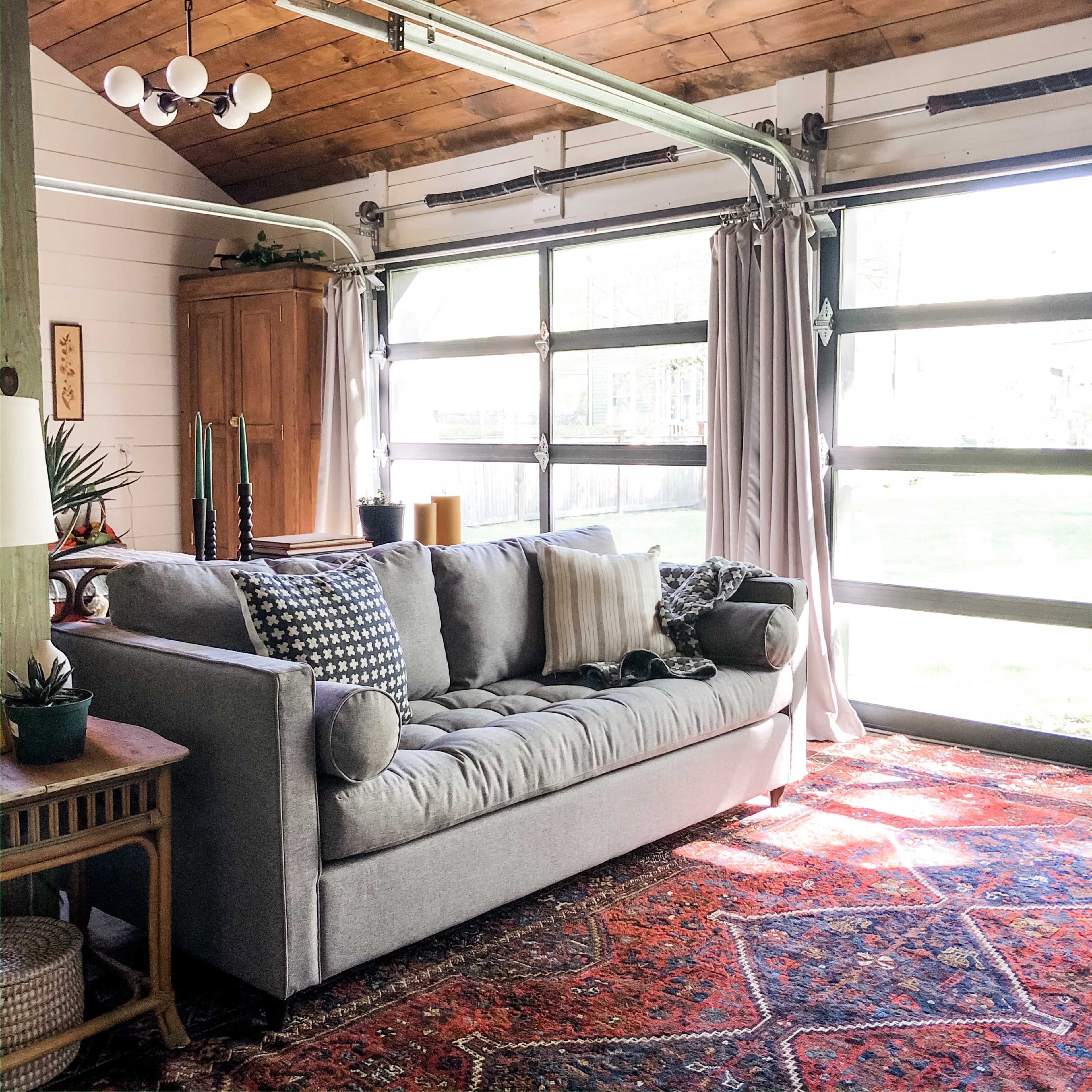 A cozy room with a gray sofa, patterned cushions, and large garage-style windows that open to a green outdoor space.