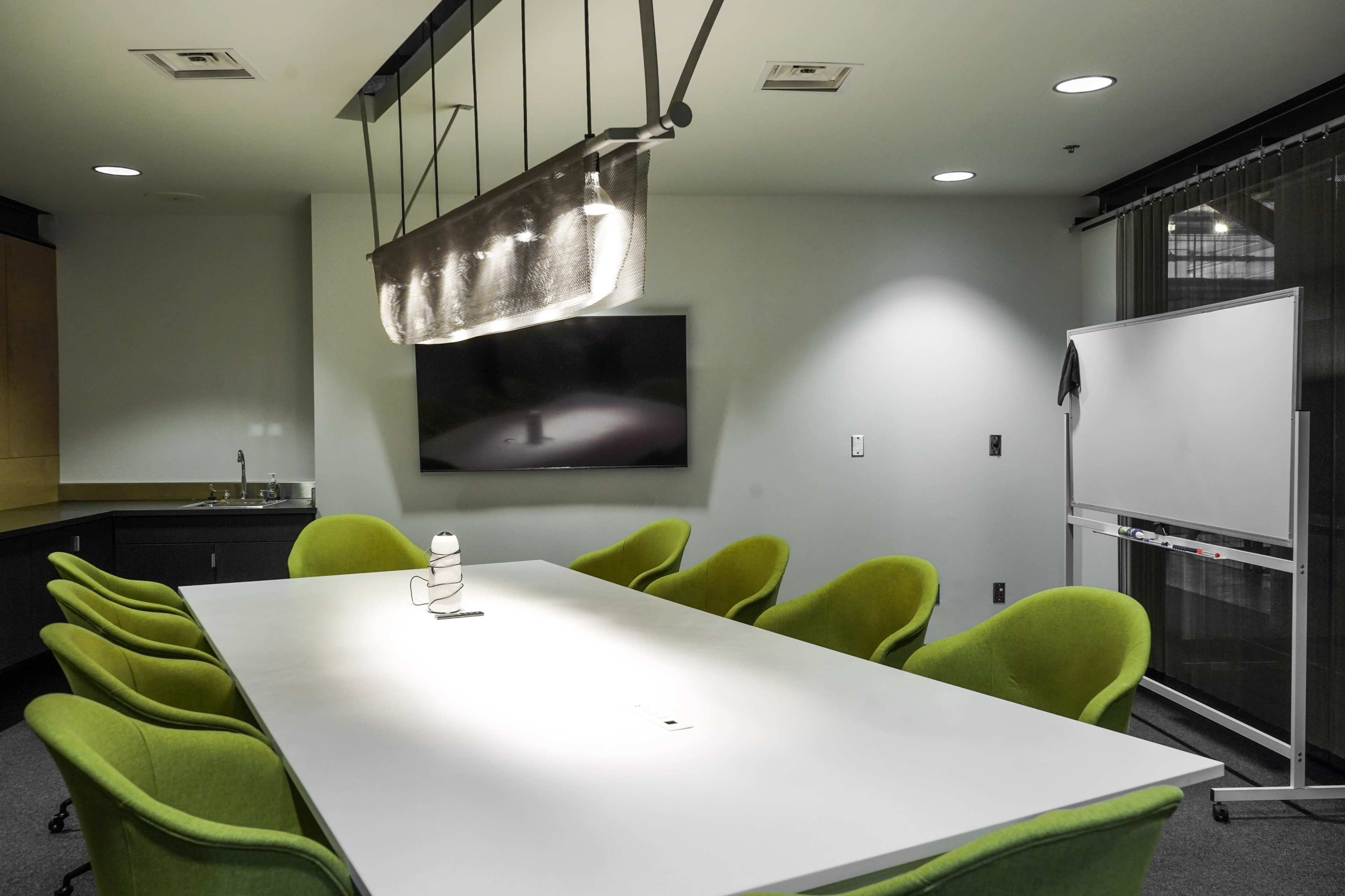 The image shows a modern meeting room featuring a large white table surrounded by green chairs, a flat-screen TV on the wall, and a whiteboard.
