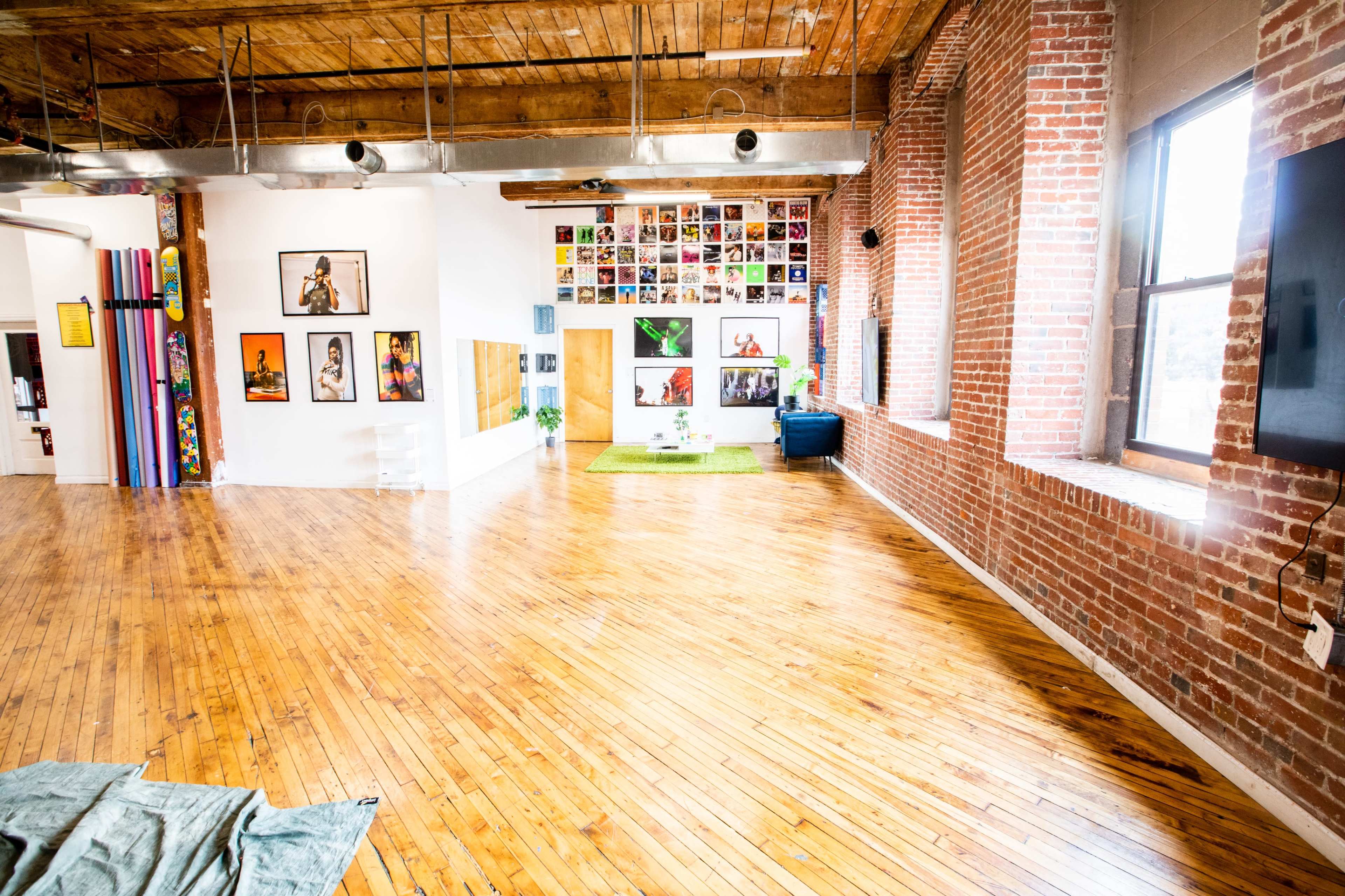 A spacious, brightly lit interior with wooden floors, brick walls decorated with framed photographs, and a small green area with plants.