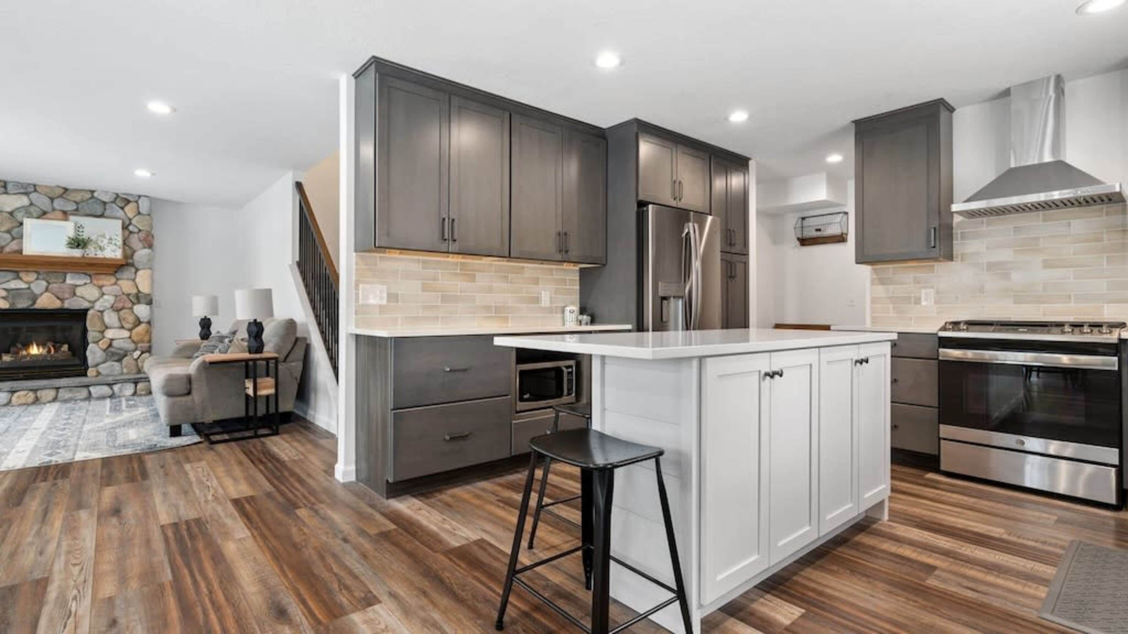 A modern kitchen with dark cabinetry, a stainless steel refrigerator, and an island with a black stool, connected to a living area featuring a stone fireplace.