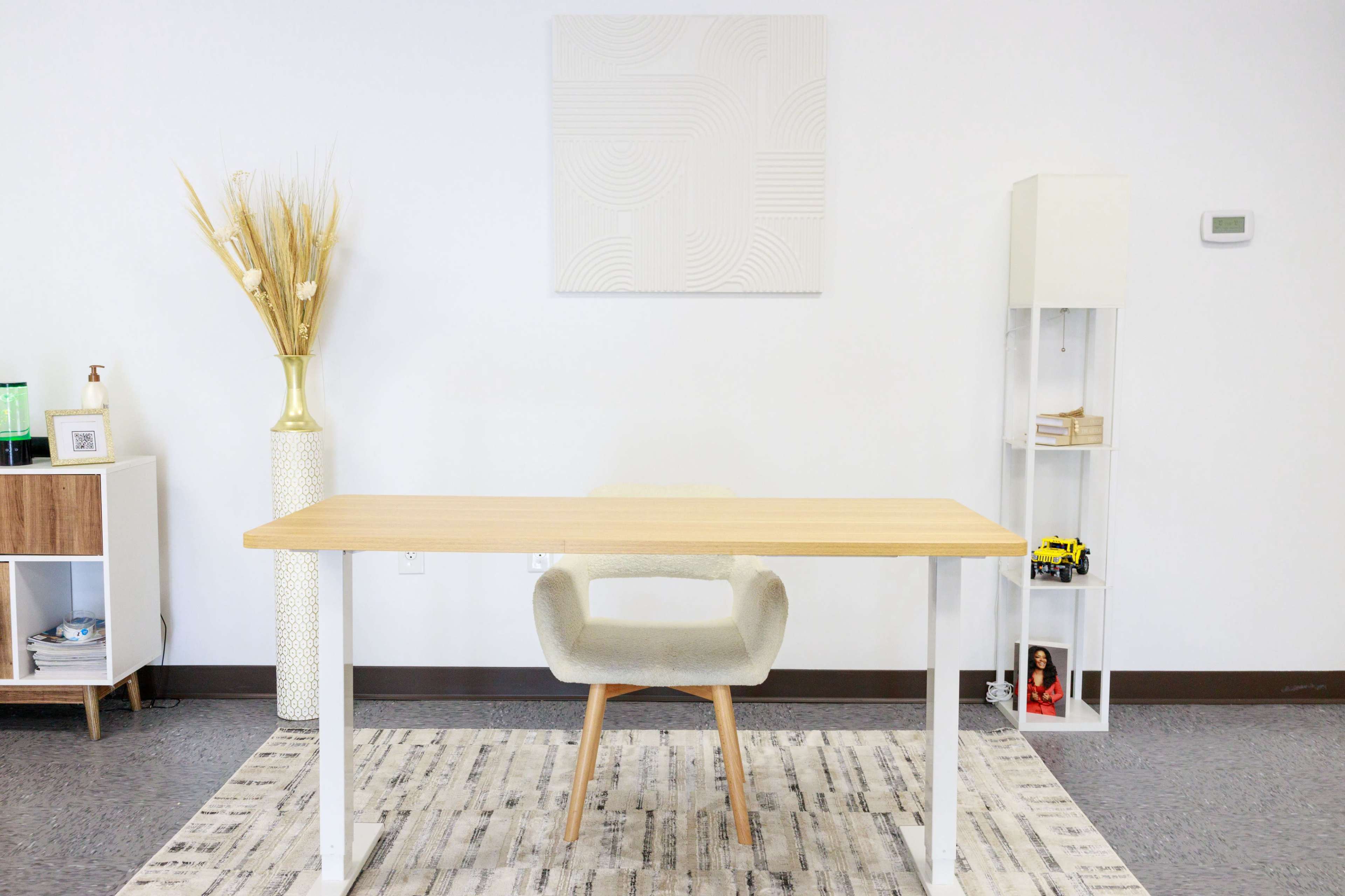A minimalist office space features a wooden desk, a light gray chair, a decorative plant in a vase, and a white shelving unit against a plain wall.