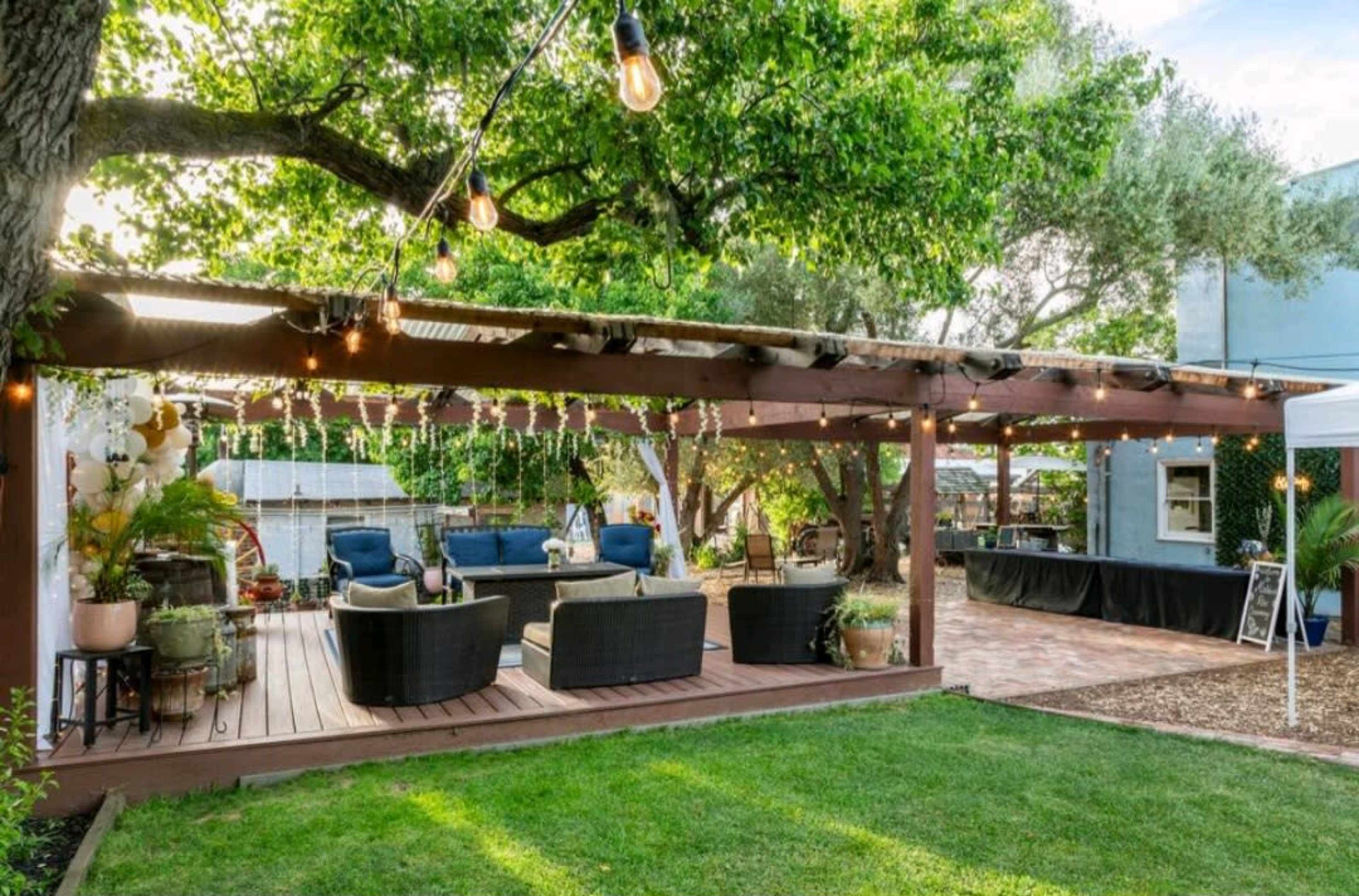 The image shows a shaded outdoor patio area with seating arranged under a wooden pergola, string lights overhead, and a grassy lawn in front.