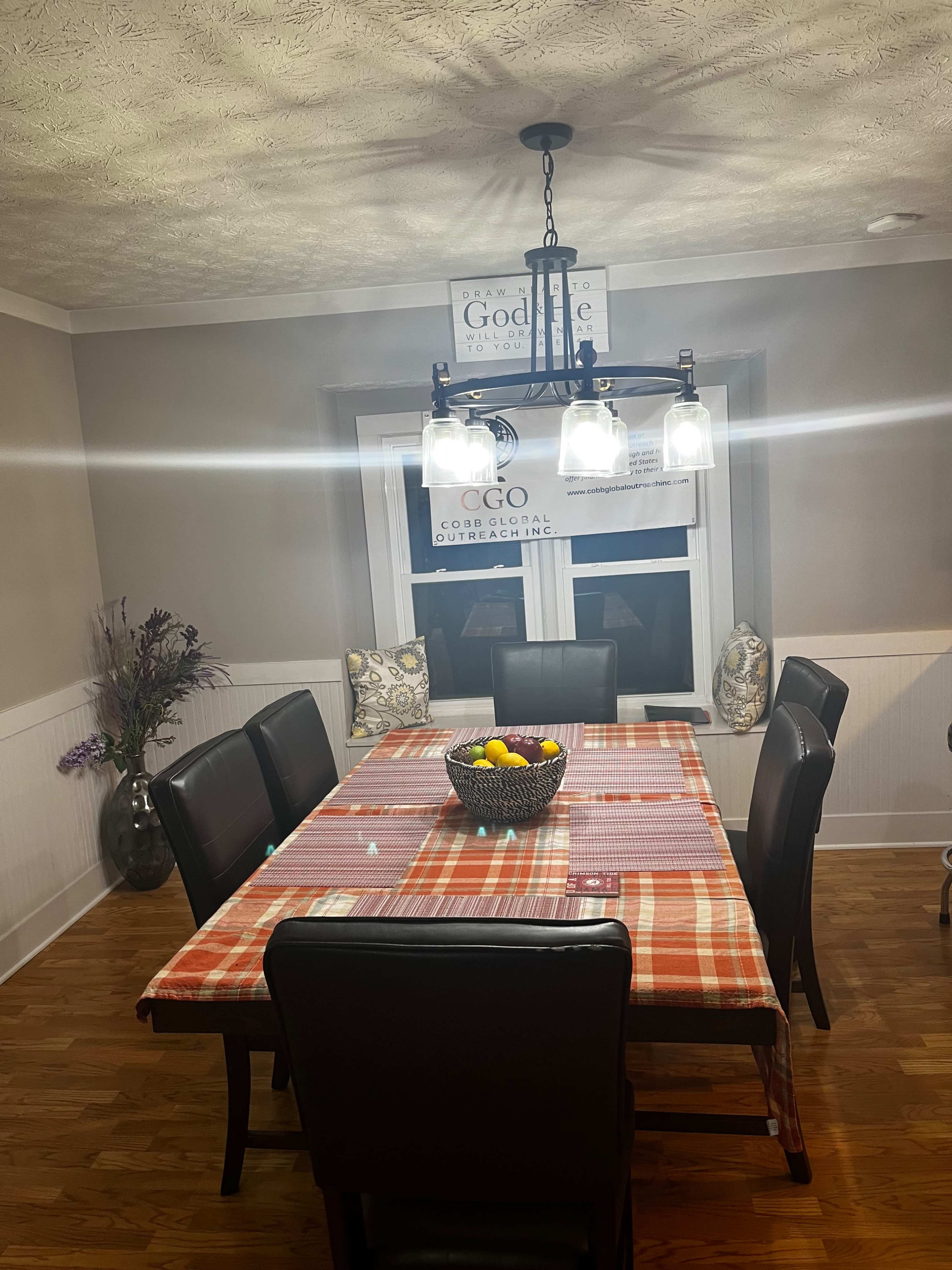 A spacious dining room features a long table set with a checkered tablecloth, surrounded by black chairs and a chandelier above, with a fruit bowl in the center.