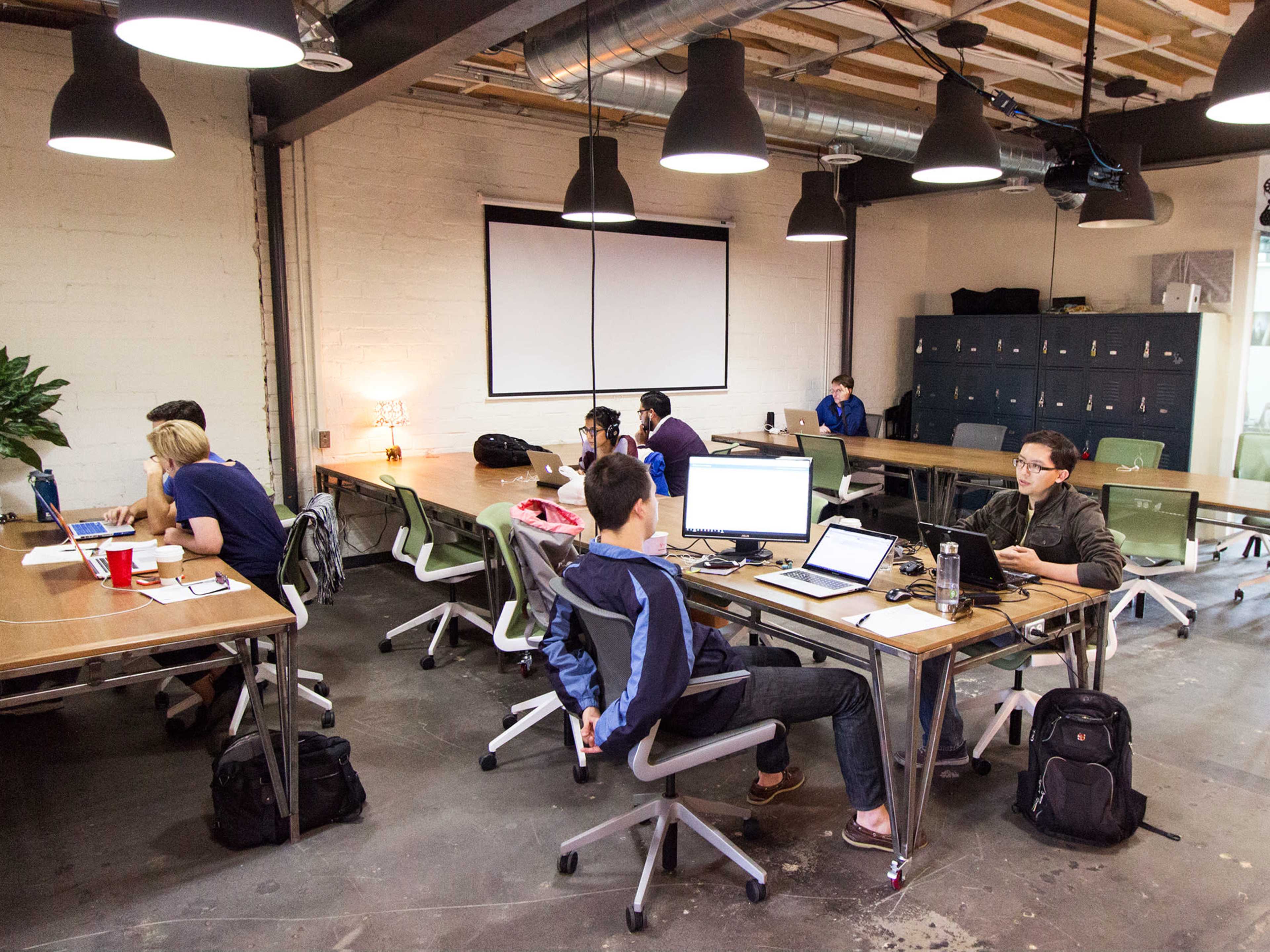 A modern office space with wooden tables and chairs, where several individuals are working on laptops and collaborating.