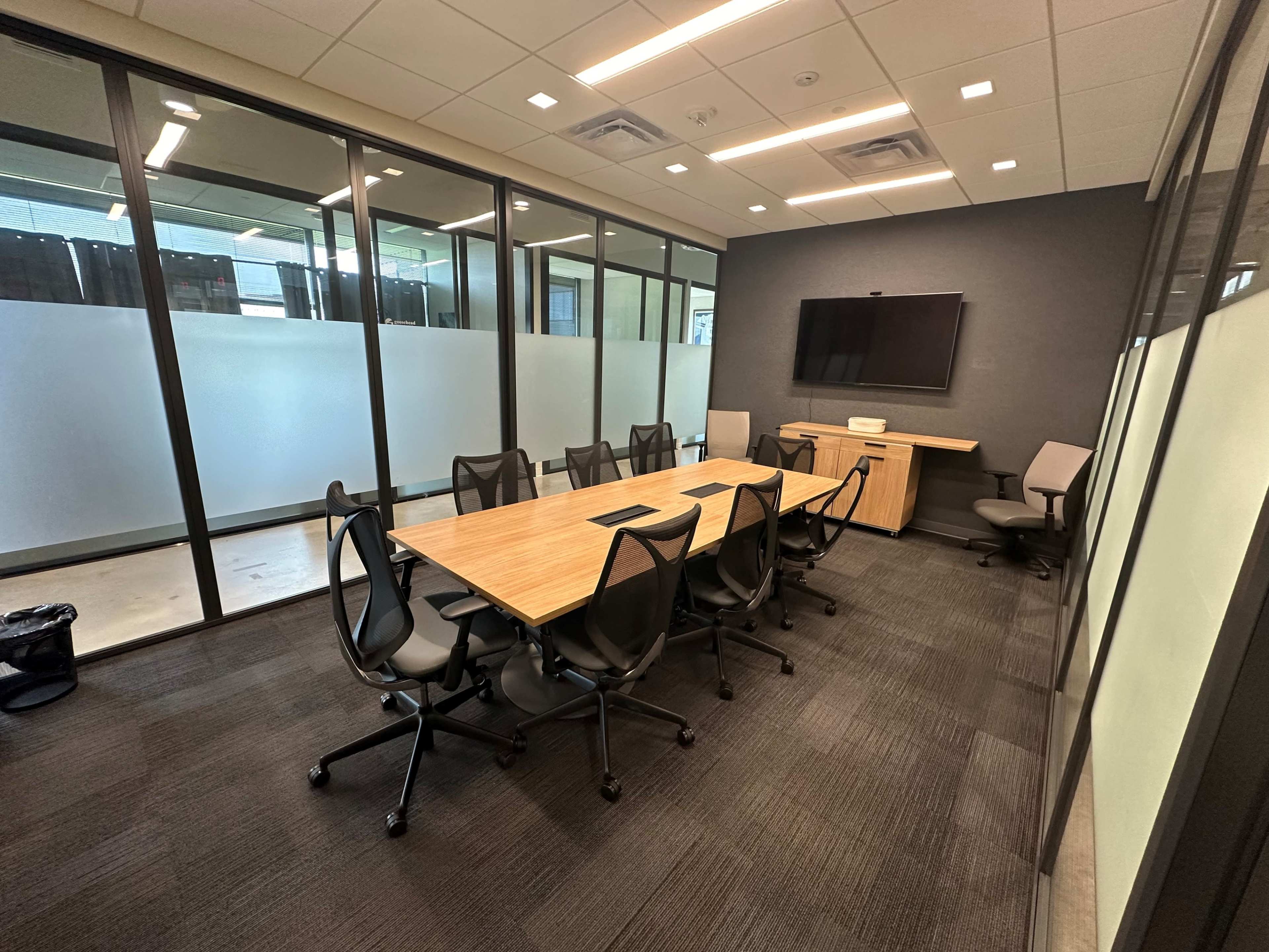 A modern conference room with a wooden table, several black chairs, and a wall-mounted television.