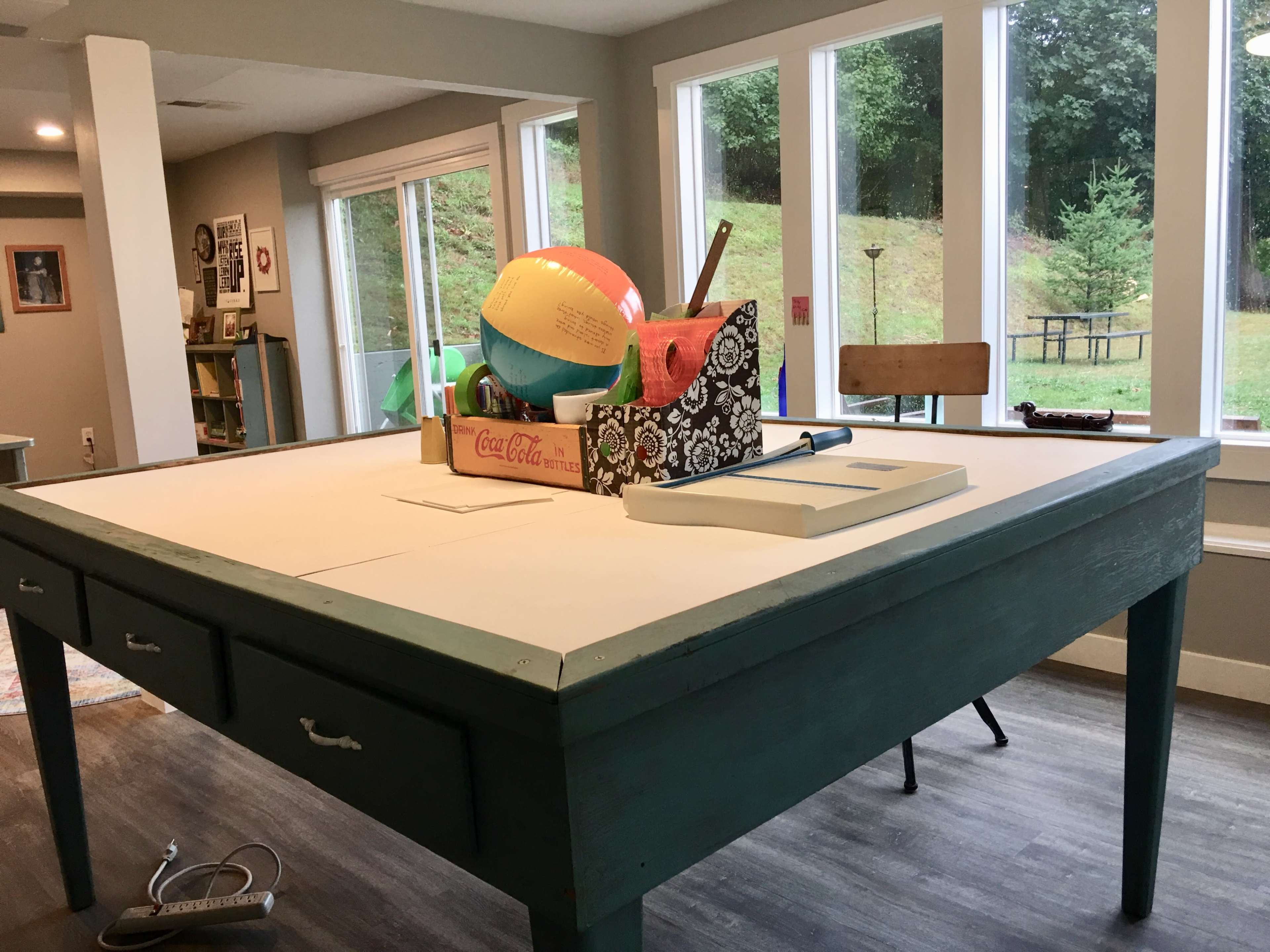 A large, painted teal table with drawers is set in a room filled with various colorful items, including a beach ball and a toolbox, while sunlight streams through the windows.