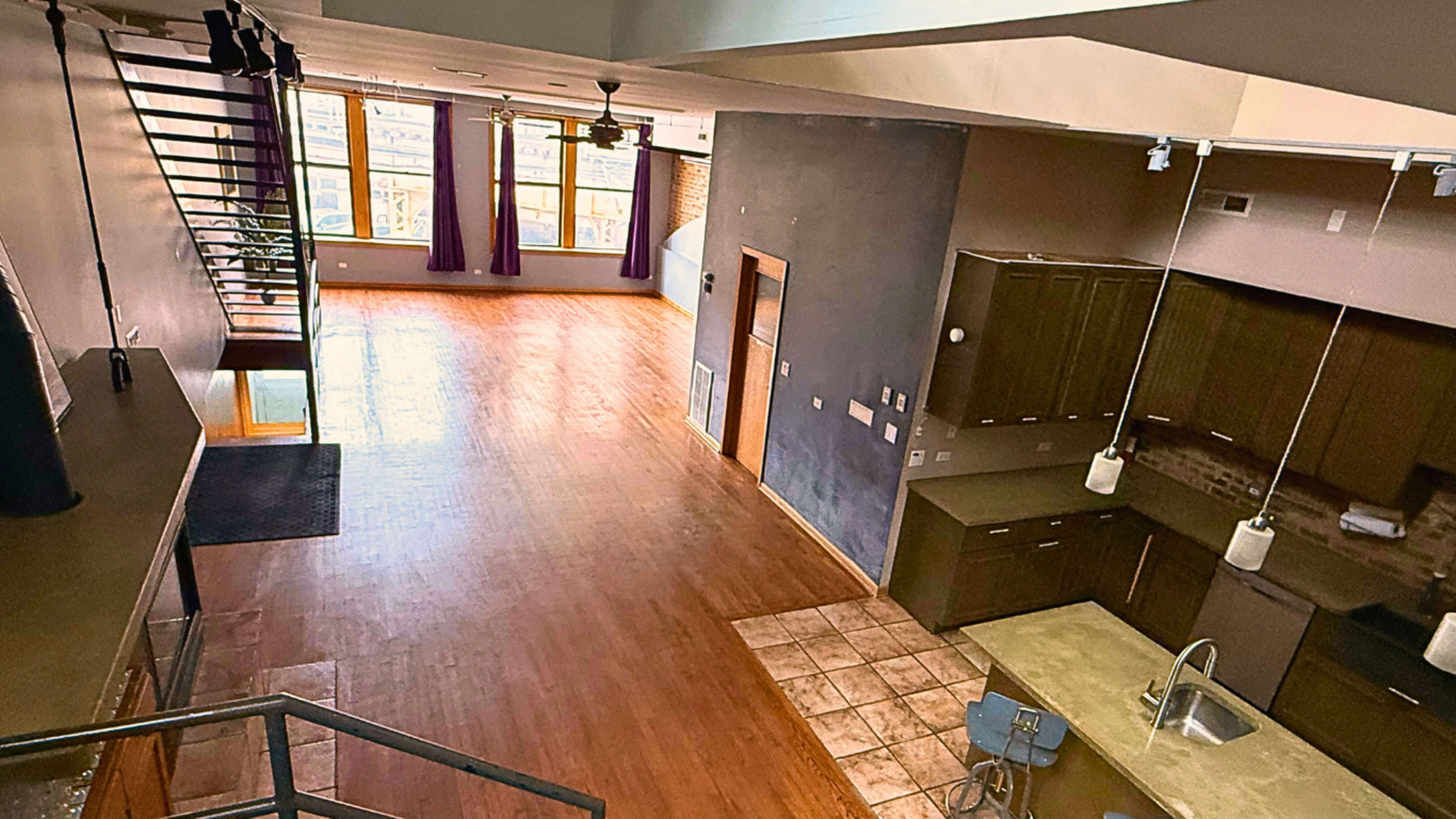 The image shows a spacious, loft-style interior featuring a modern kitchen, an open living area with large windows, and a staircase leading to an upper level.