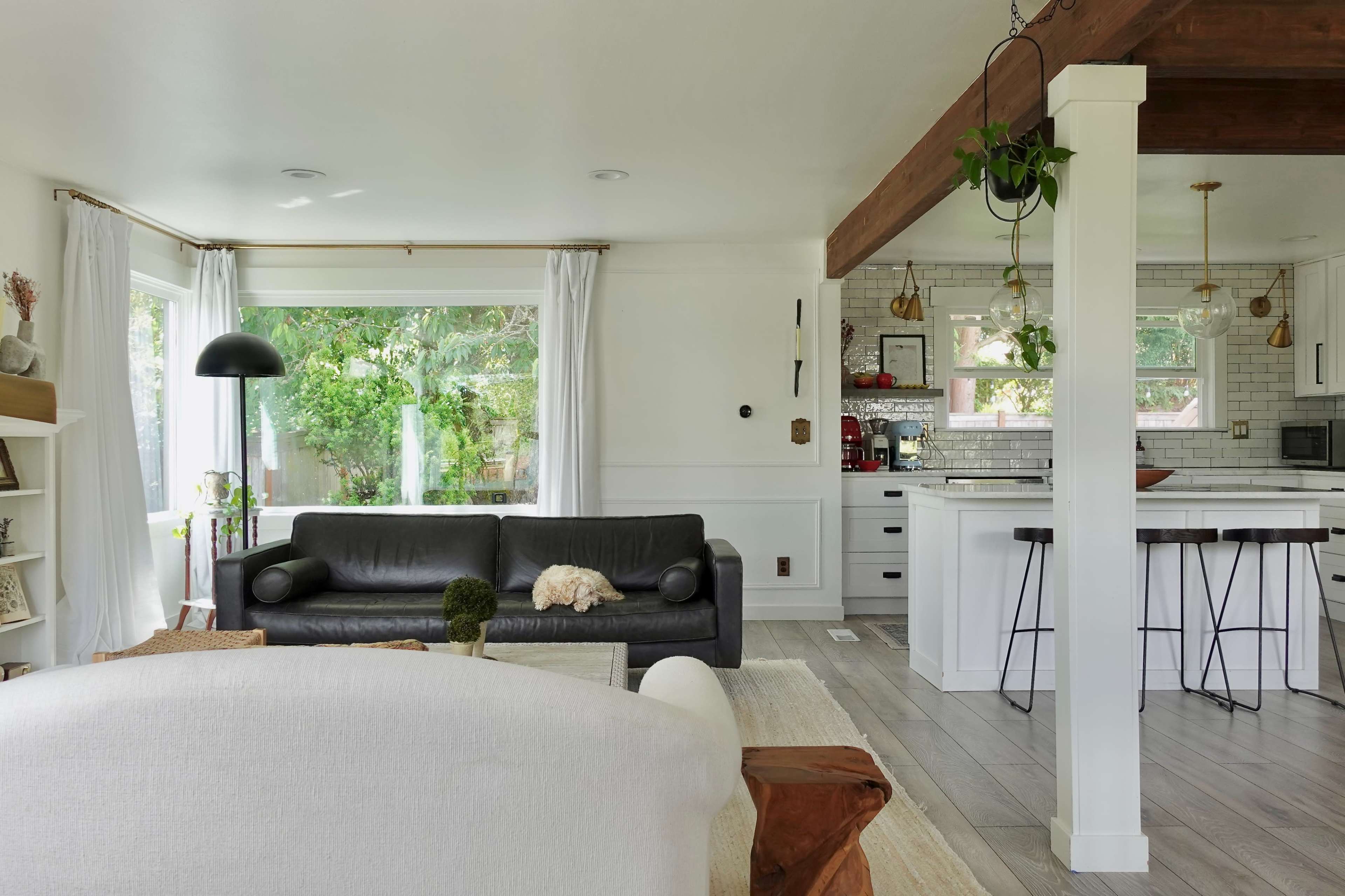 A modern living space featuring a black sofa with a dog resting on it, a light area rug, and a kitchen area with bar stools and white cabinetry.