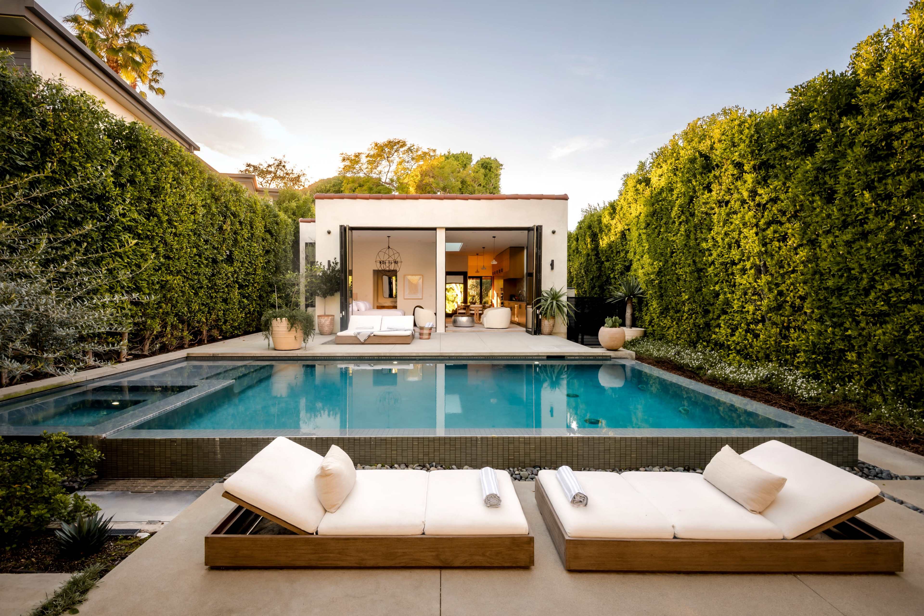 The image features a modern backyard with a swimming pool, lounge chairs, and a landscaped area surrounded by tall greenery.