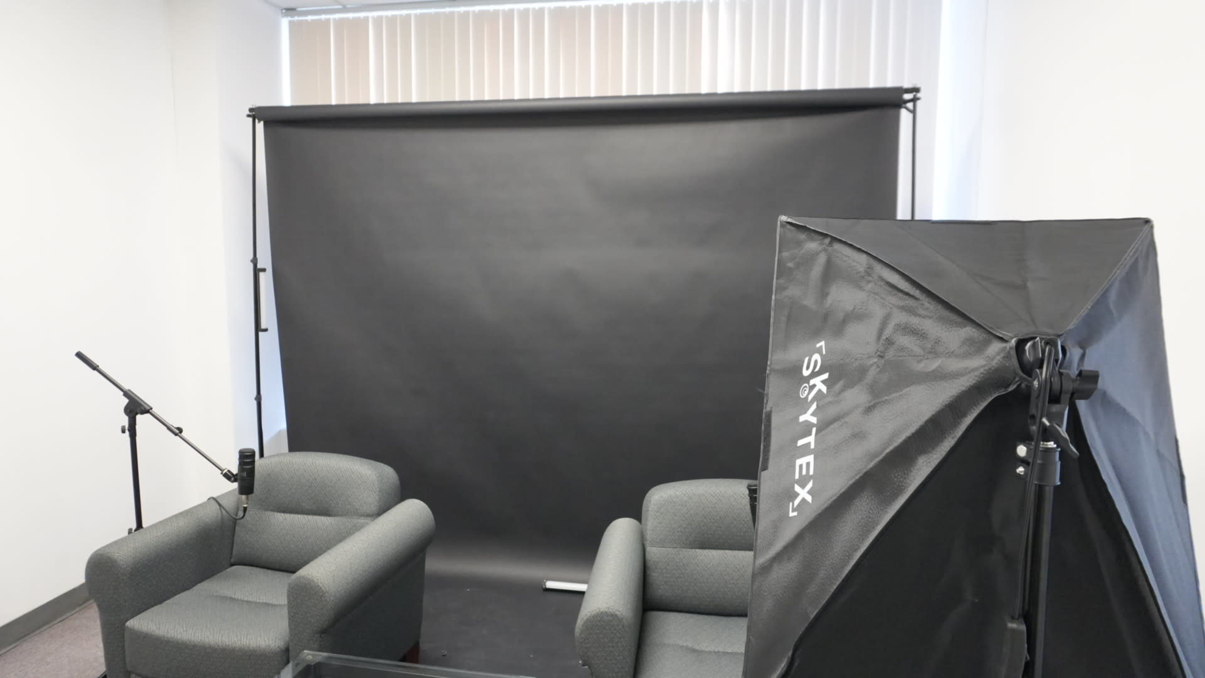 The image shows a small studio setup with two gray chairs, a black backdrop, and a softbox light positioned nearby.