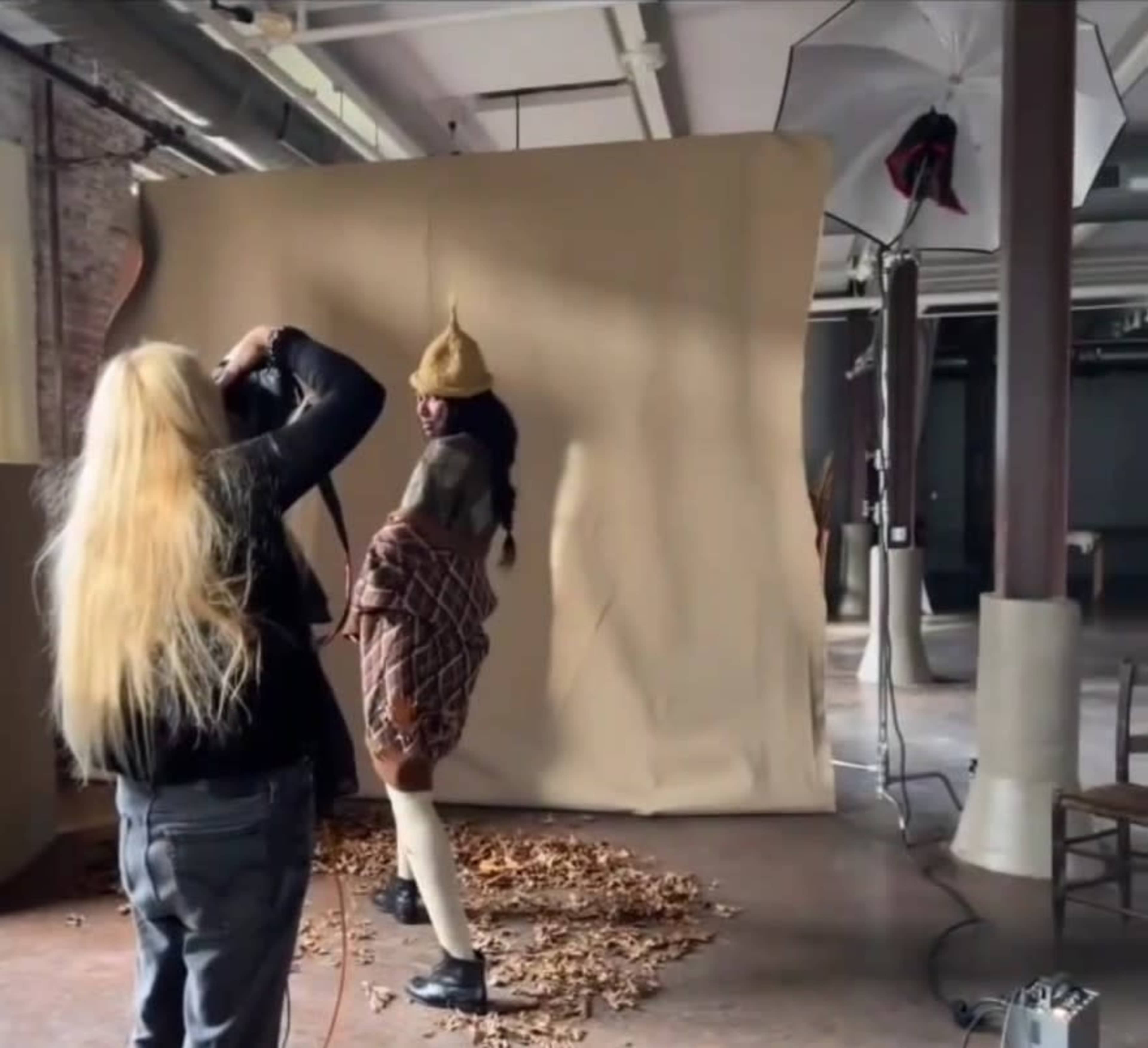 A photographer captures a model posing in front of a beige backdrop scattered with autumn leaves.