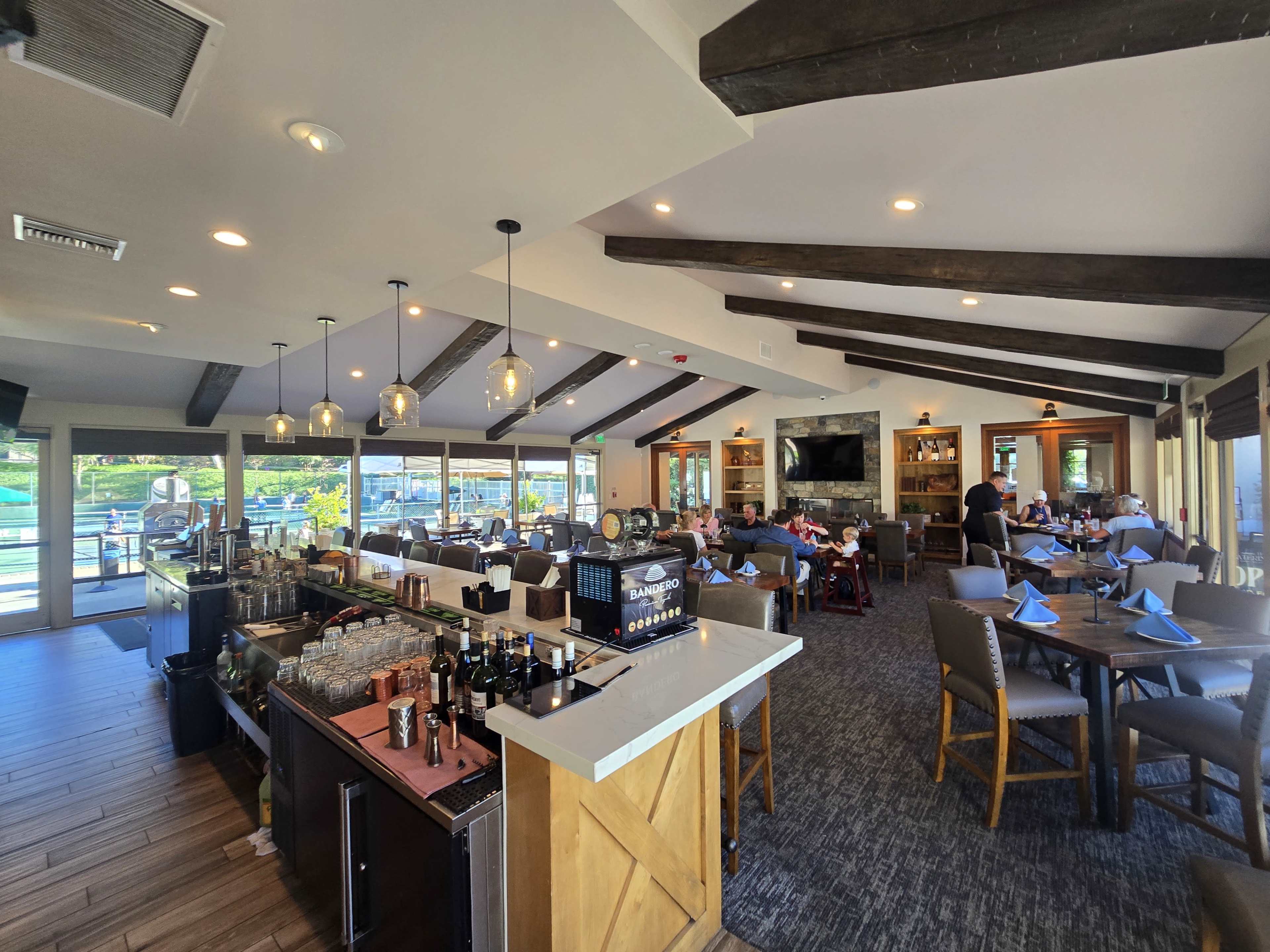 The image shows the interior of a restaurant featuring a bar area with glasses and bottles, dining tables with patrons, and large windows that offer views of the outside.