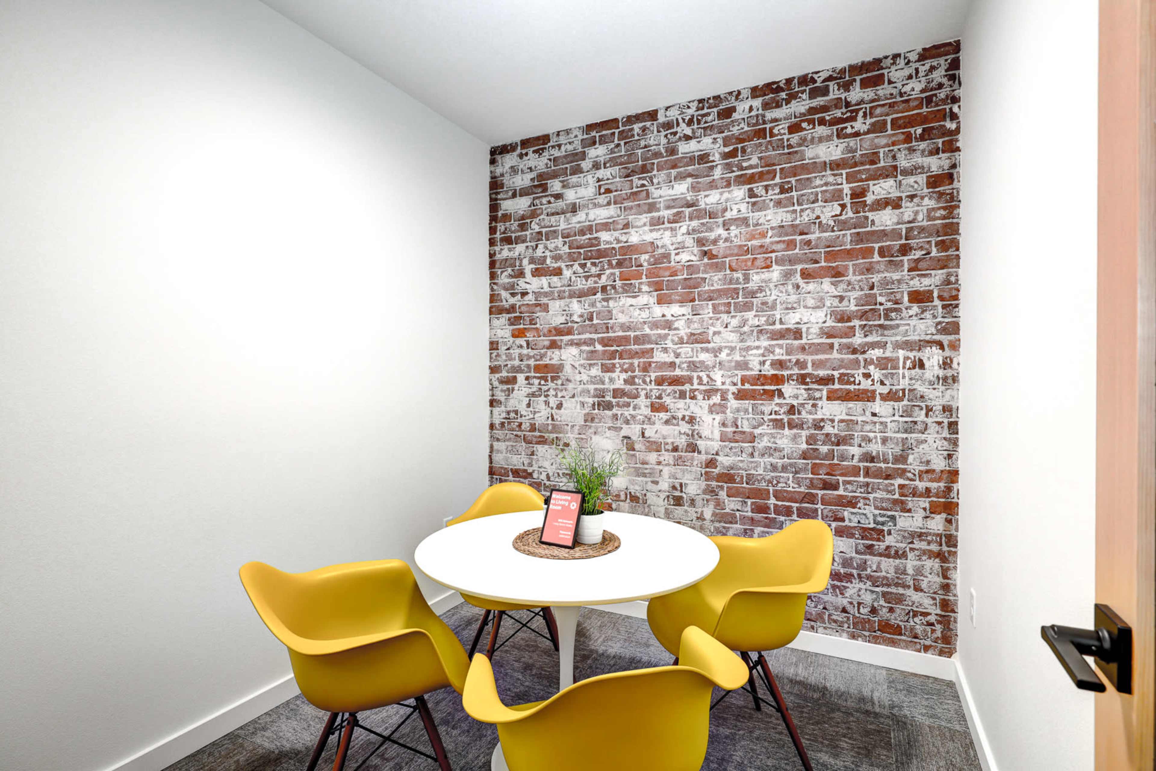 A small meeting room features a round white table surrounded by four yellow chairs and a red brick accent wall.