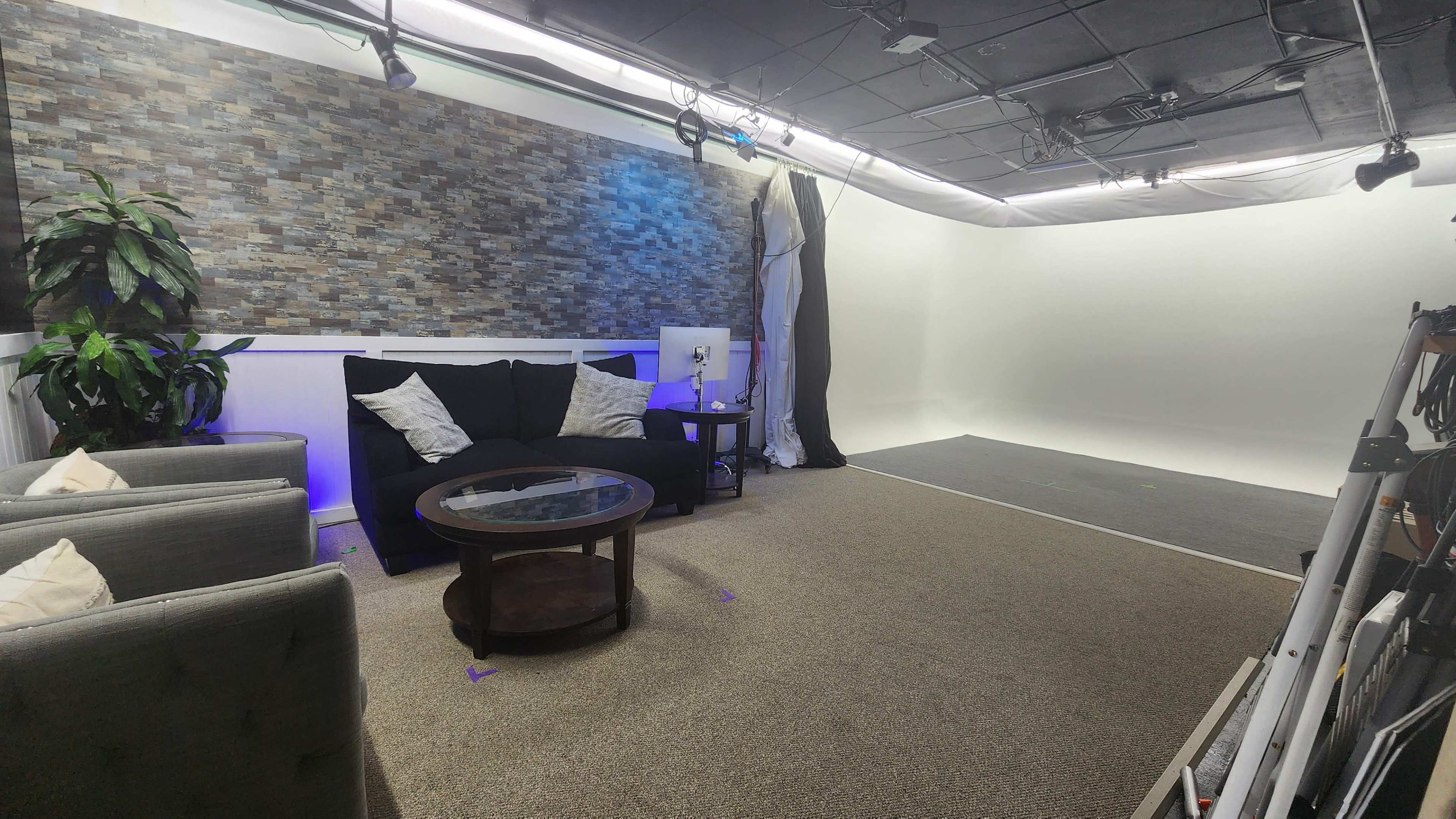A modern studio setup with a textured stone wall, a black sofa with decorative pillows, a glass coffee table, and a large empty white backdrop.