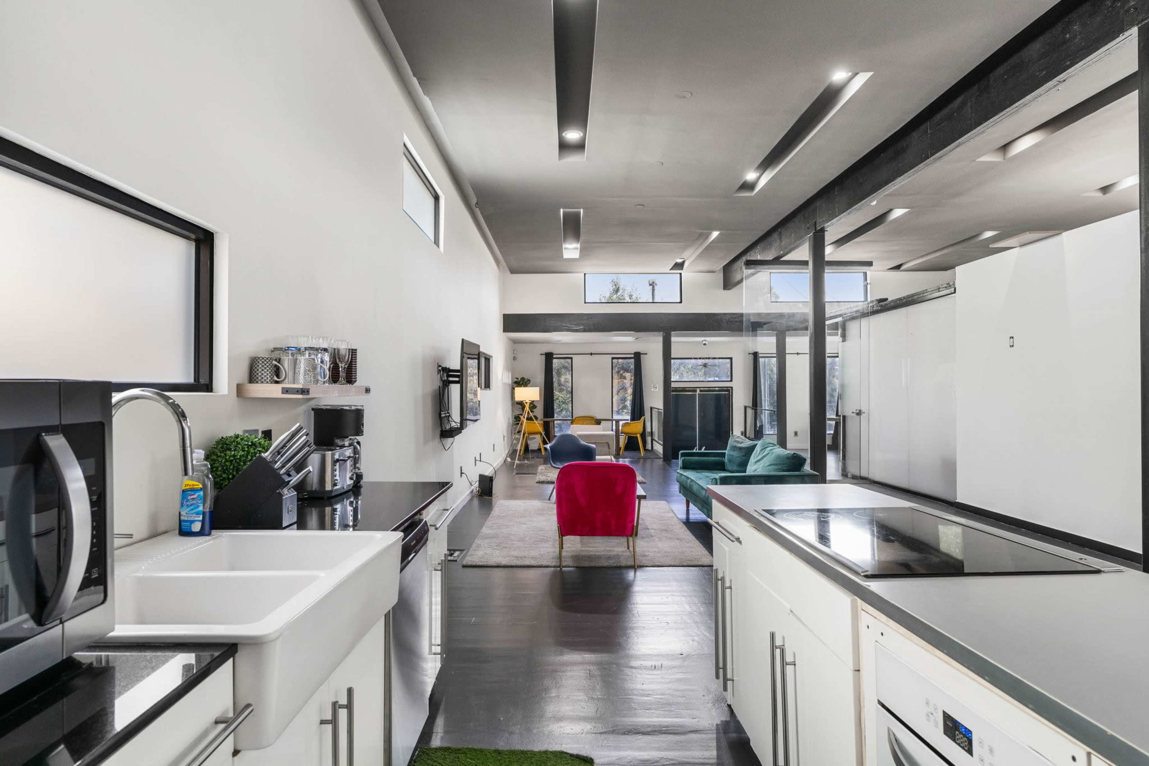 A modern kitchen with a sink, microwave, and cooking area opens to a spacious living area featuring colorful furniture and large windows.