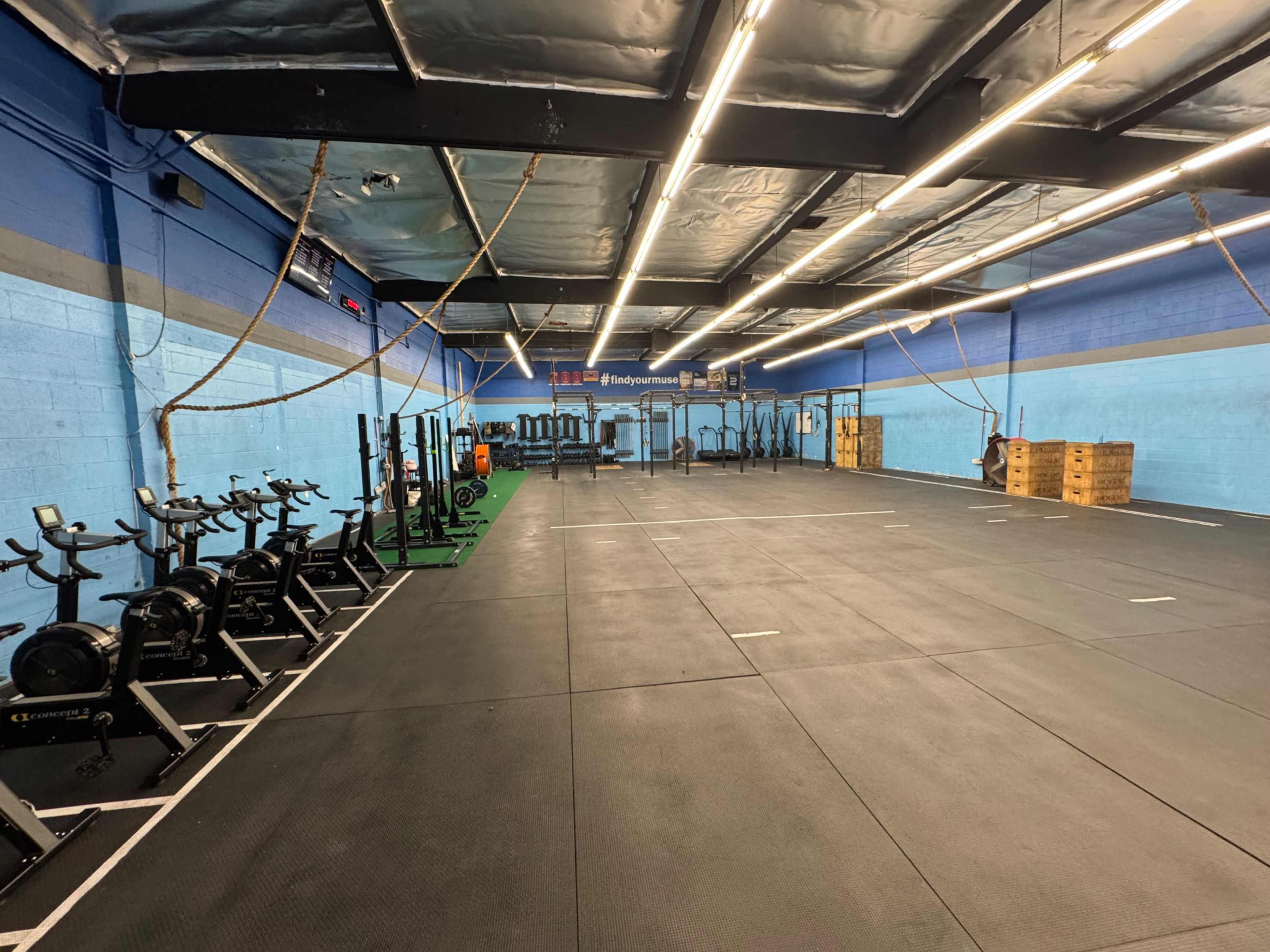 The image shows a spacious gym interior with exercise equipment, including rowing machines and weightlifting stations, arranged neatly on a black rubber floor.