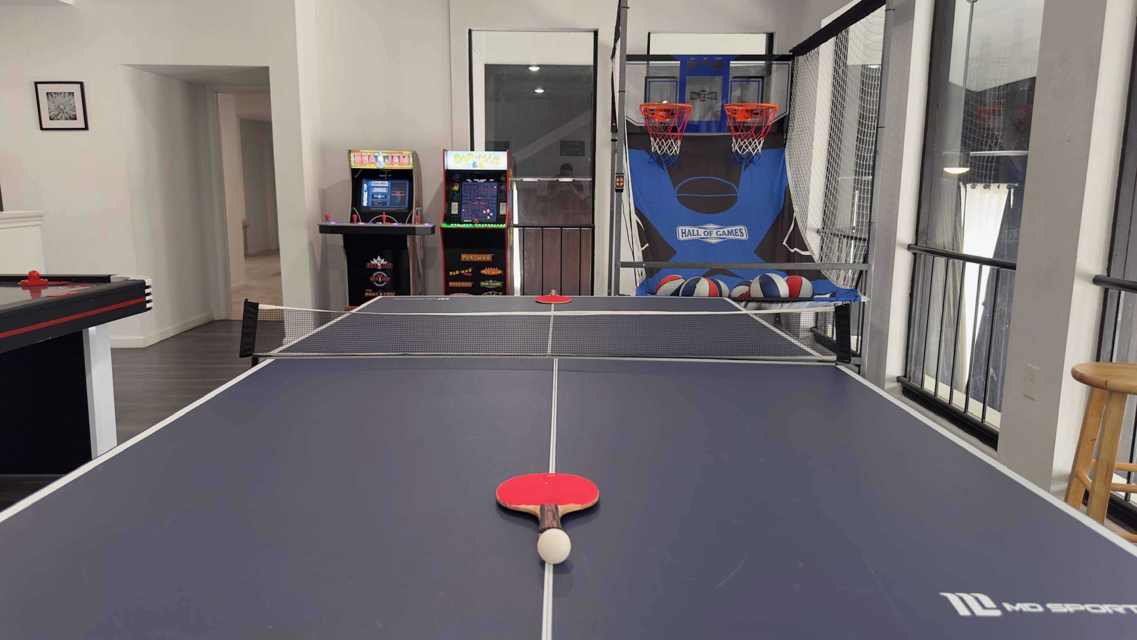 A game room featuring a table tennis table with a paddle and ball in the foreground, alongside arcade games and a basketball shooting game in the background.