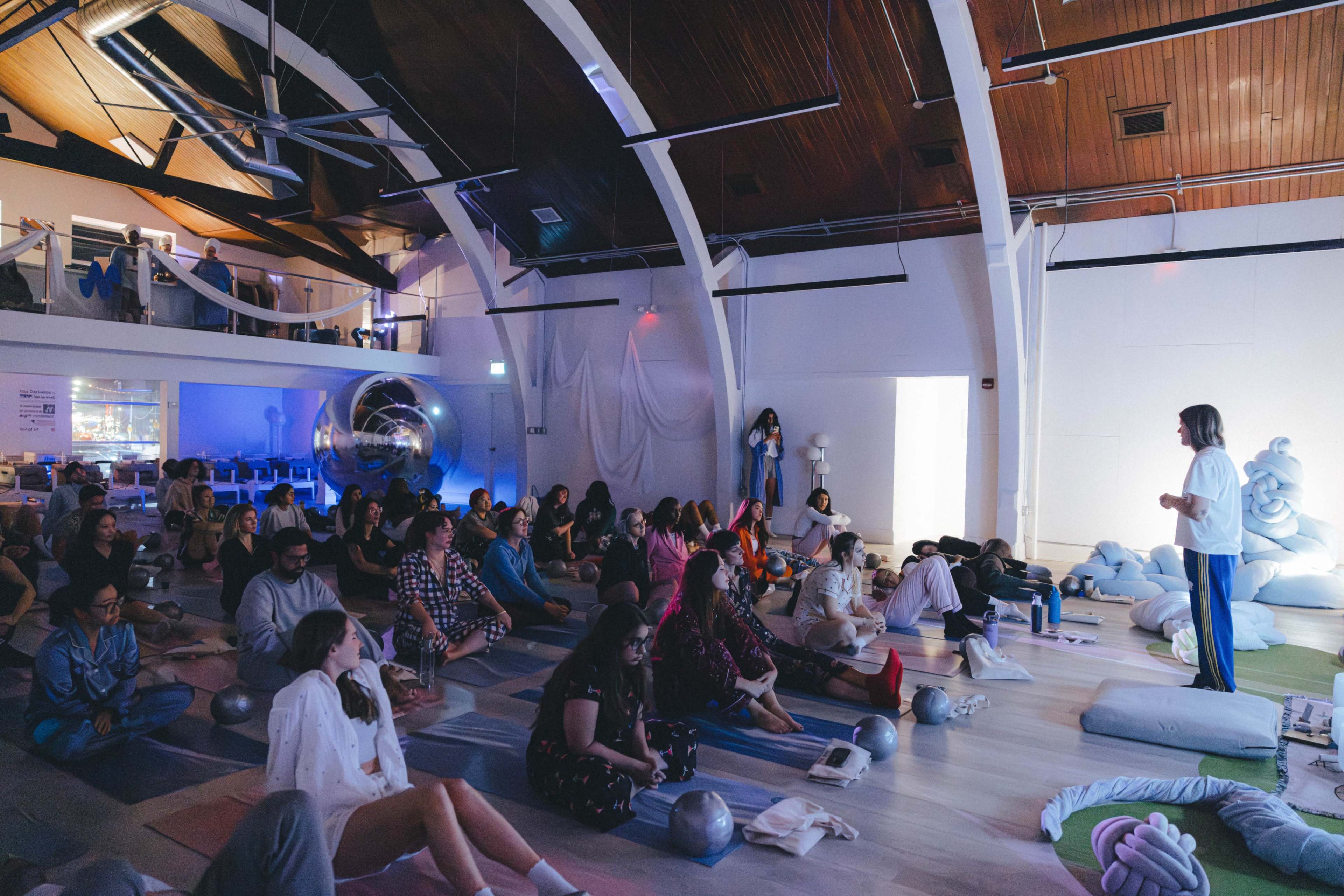 A diverse group of people sits on mats and cushions in a brightly lit space while a person speaks to them.