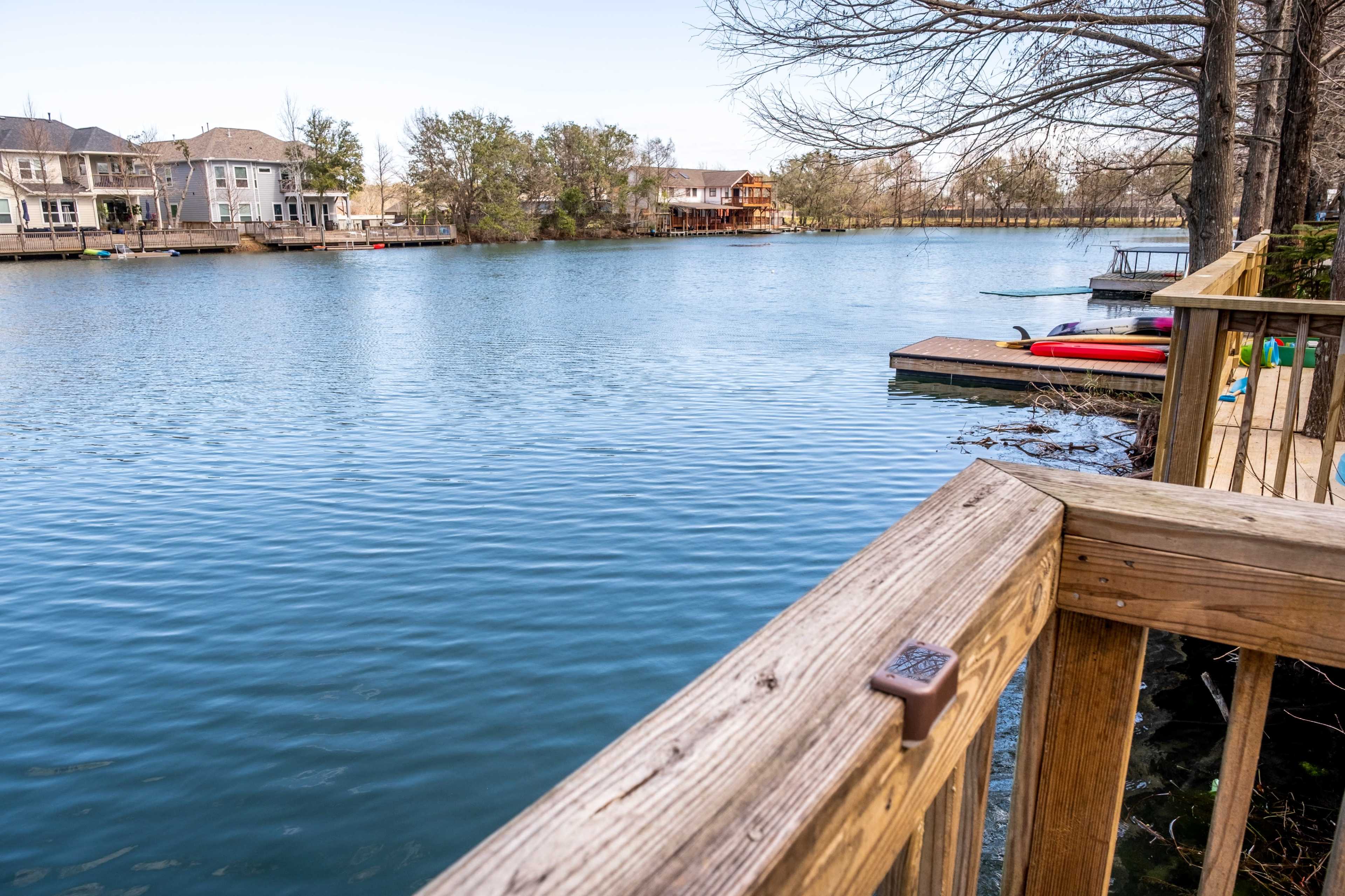 A calm lake is lined with houses on one side and a wooden dock on the other, with several kayaks and paddleboards visible.