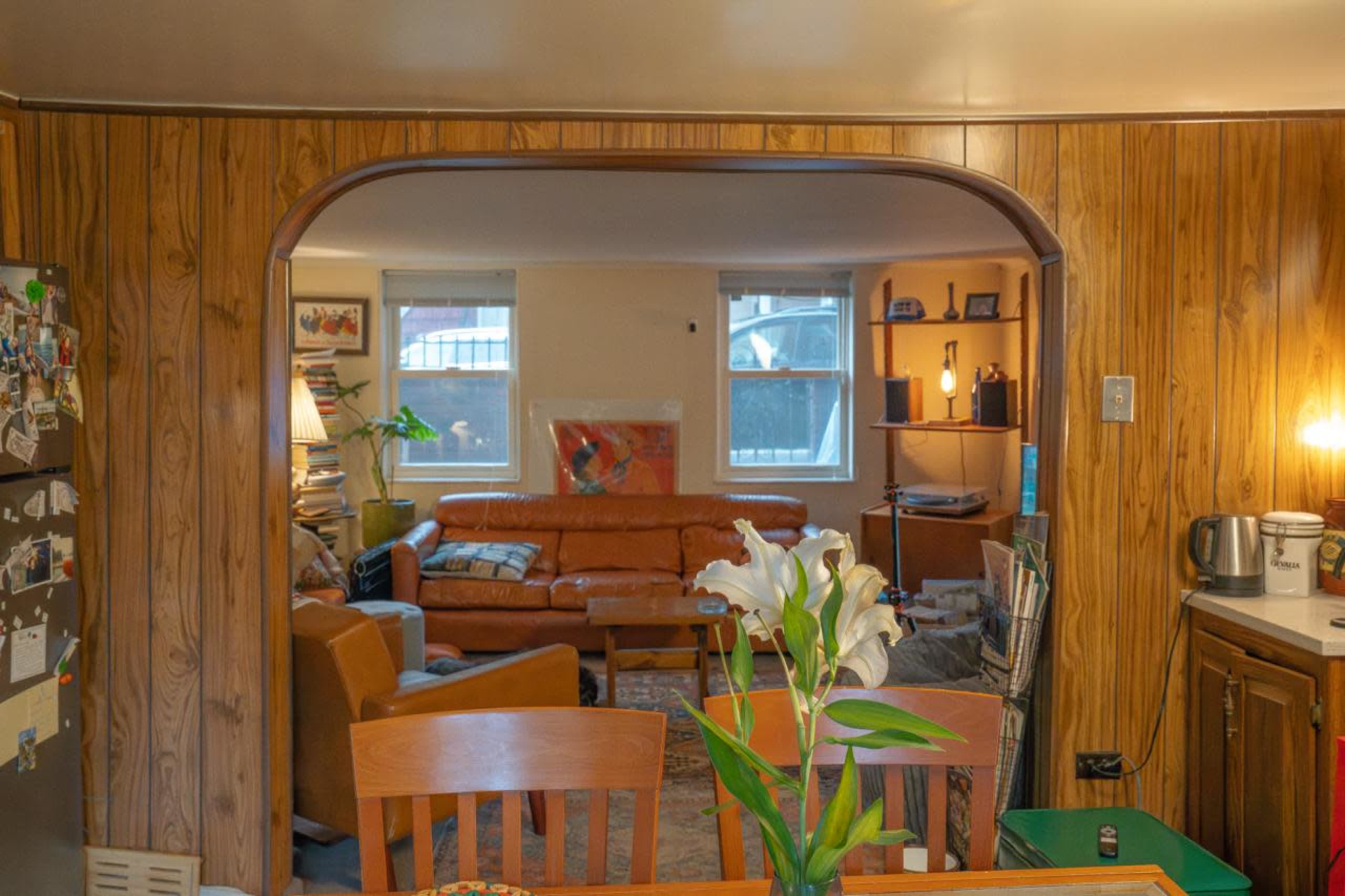 The image shows a cozy interior space featuring a kitchen area that opens up to a living room with a brown leather sofa and wooden accents.