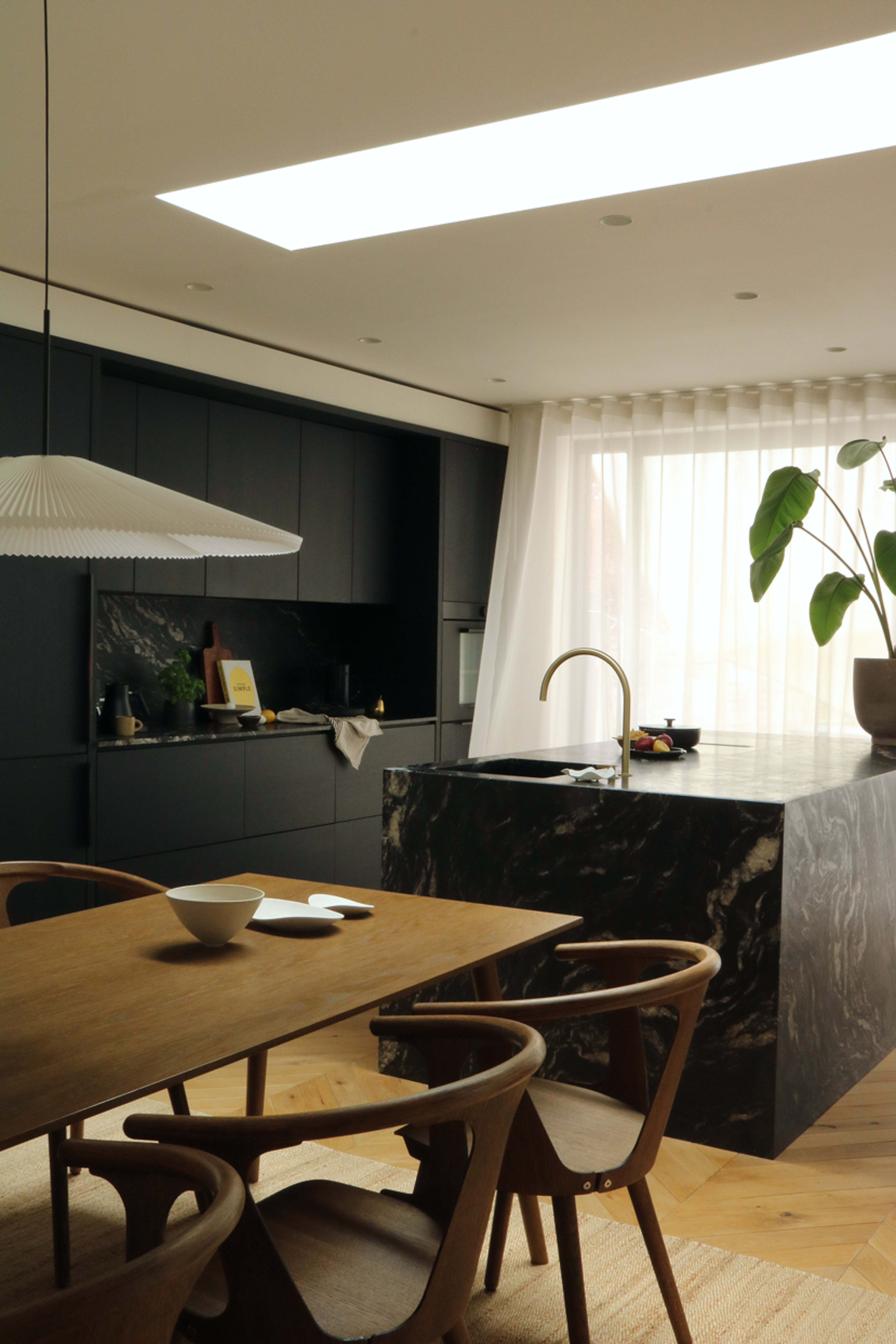A modern kitchen features a marble island and dark cabinetry, with a dining table and stylish chairs positioned nearby under a hanging light.