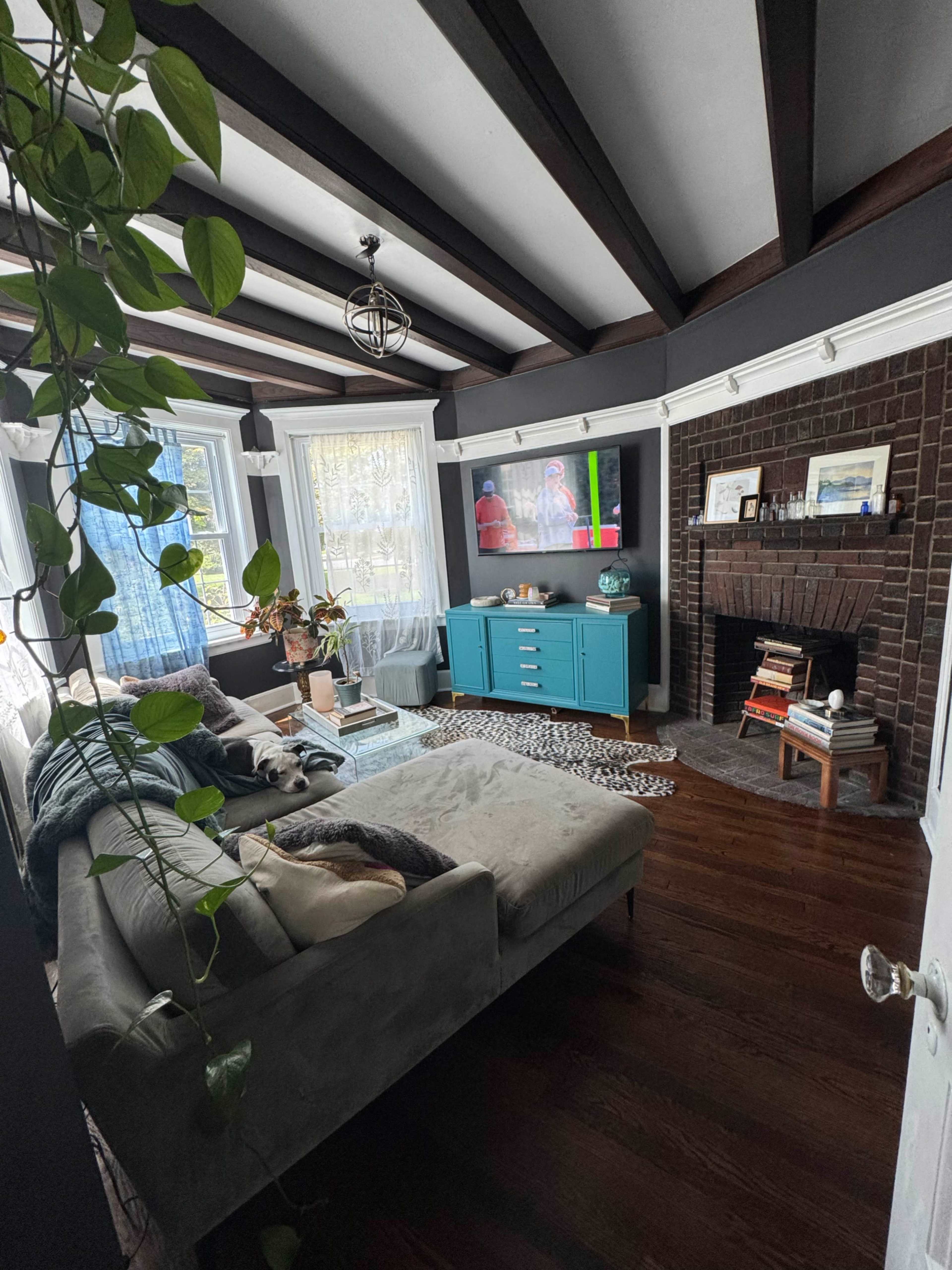 The image shows a cozy living room with a sectional sofa, a turquoise cabinet, a television mounted on the wall, and a fireplace on the right.