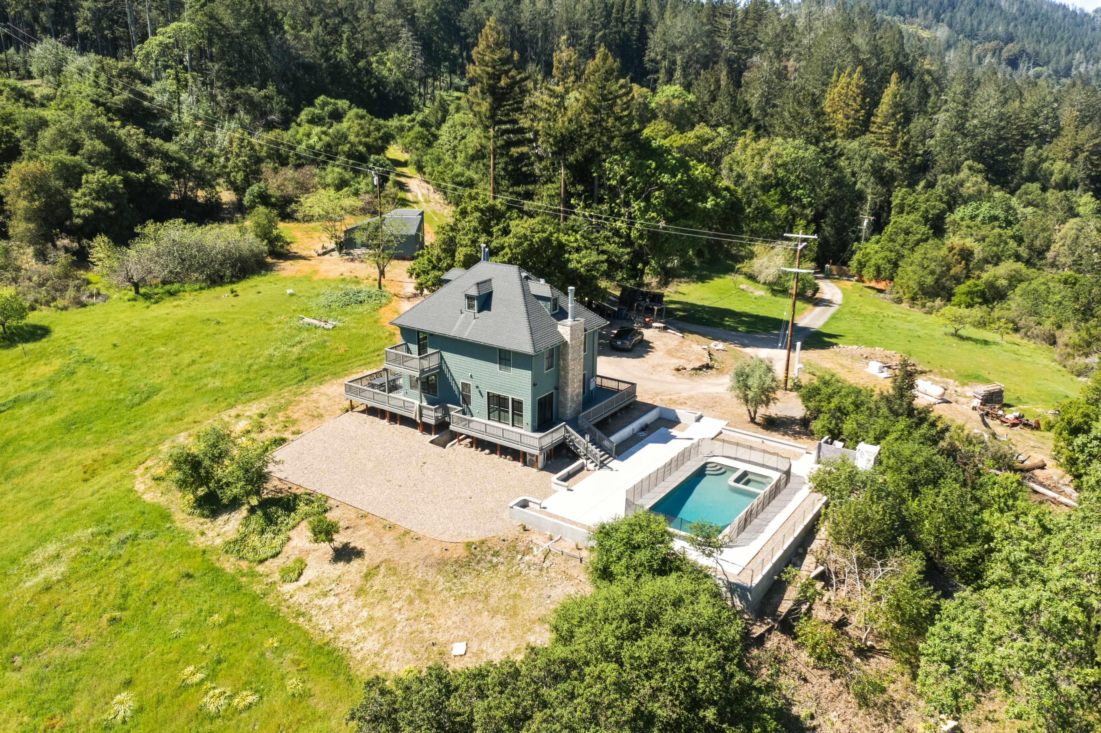 A two-story green house with a pool is situated on a grassy lot surrounded by trees.