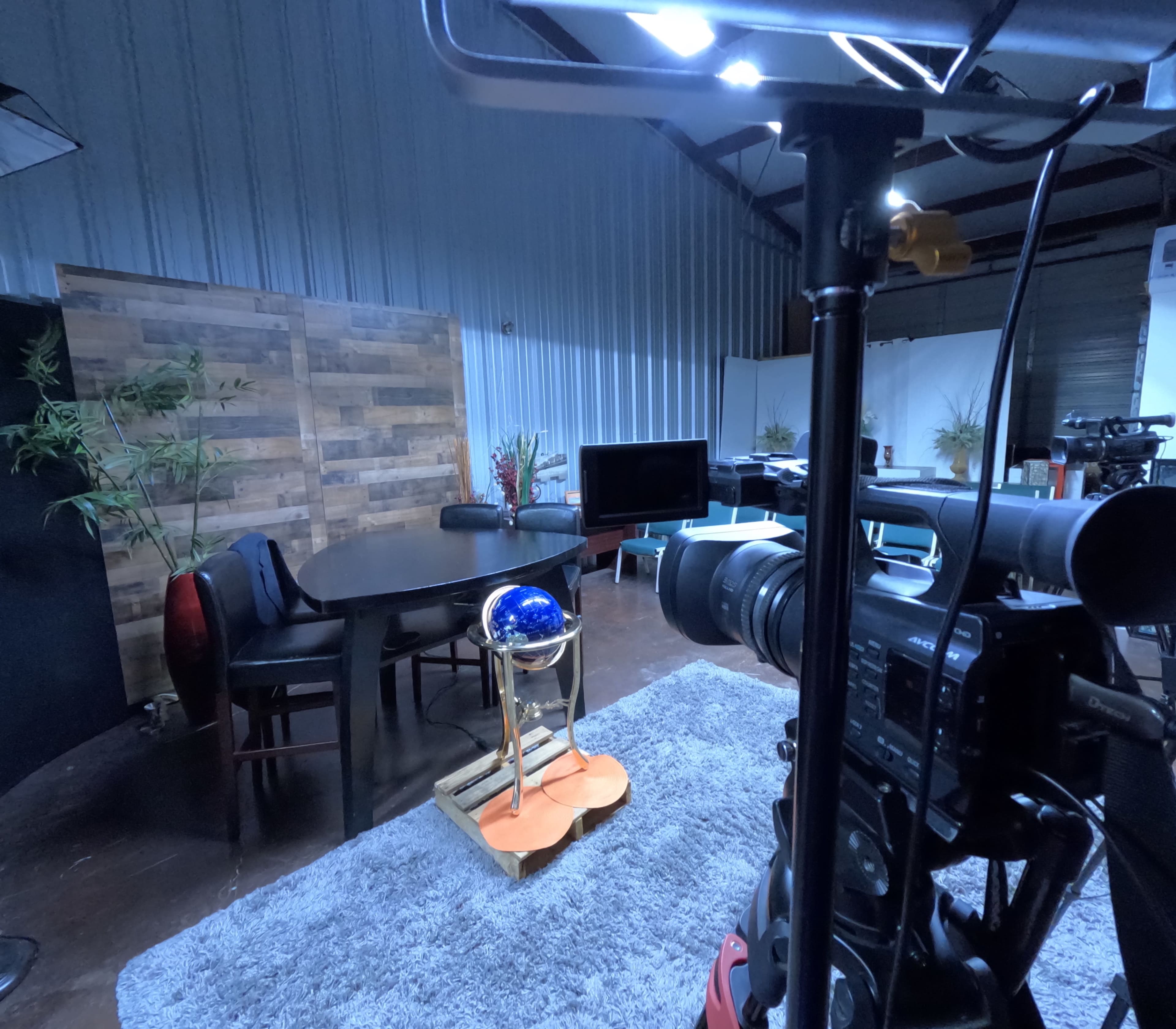 The image shows a studio setup with a round table, chairs, and a globe on a wooden stand, surrounded by cameras and decorative plants.