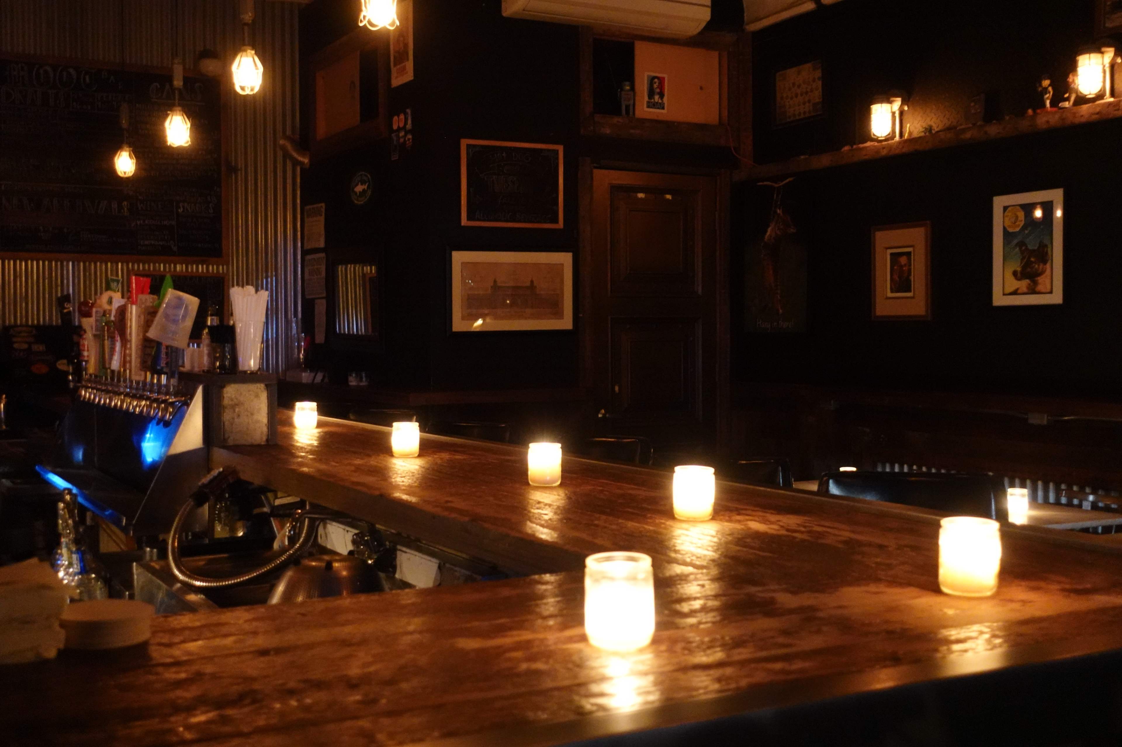 A dimly lit bar with candles arranged along the wooden counter and framed artwork on the walls.