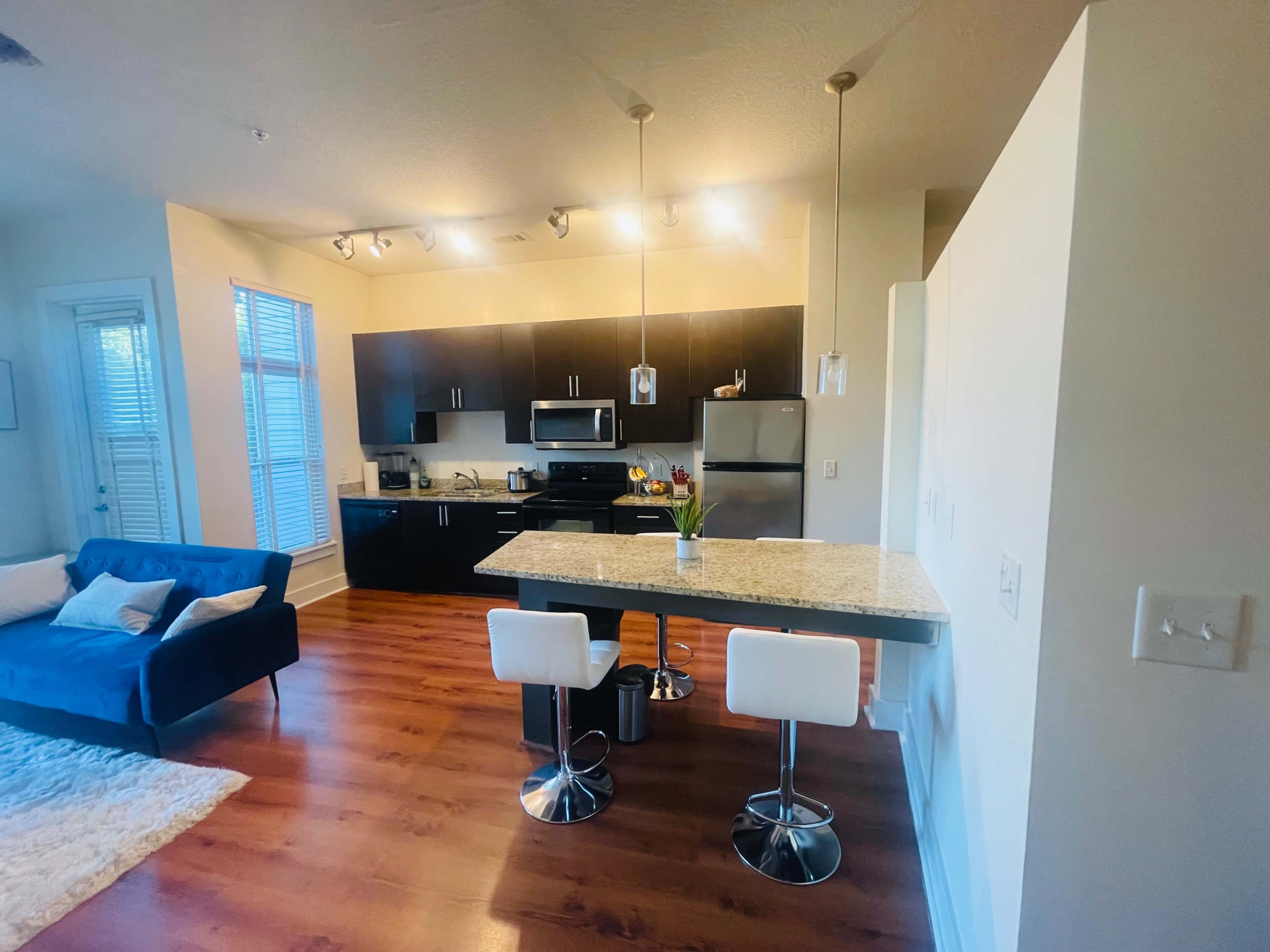 A modern kitchen and dining area with dark cabinetry, granite countertops, a small dining table with two white bar stools, and a blue sofa in the living space.
