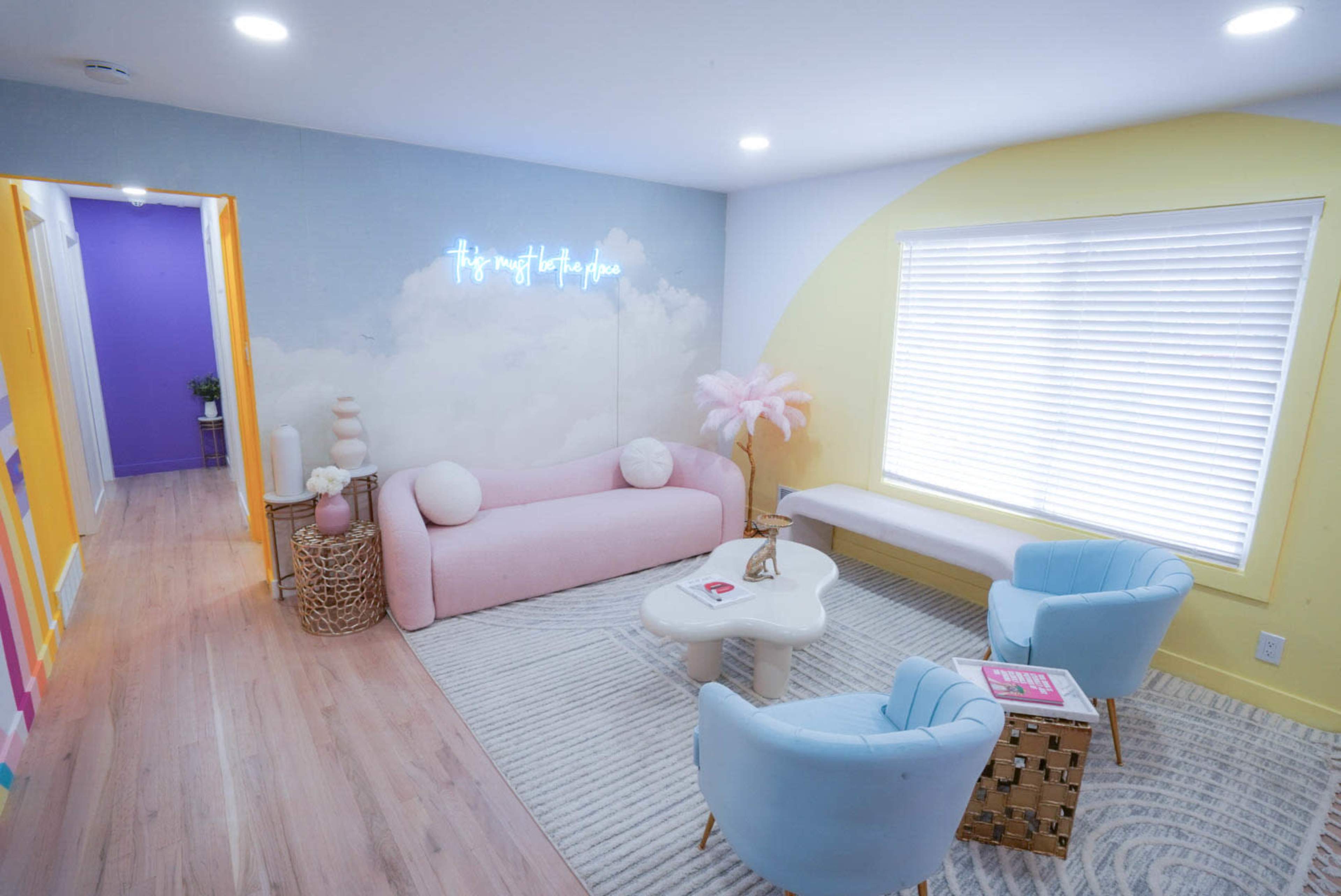 A brightly colored living room featuring a pink and white sofa, two light blue chairs, and a wall adorned with a neon sign and a cloud mural.