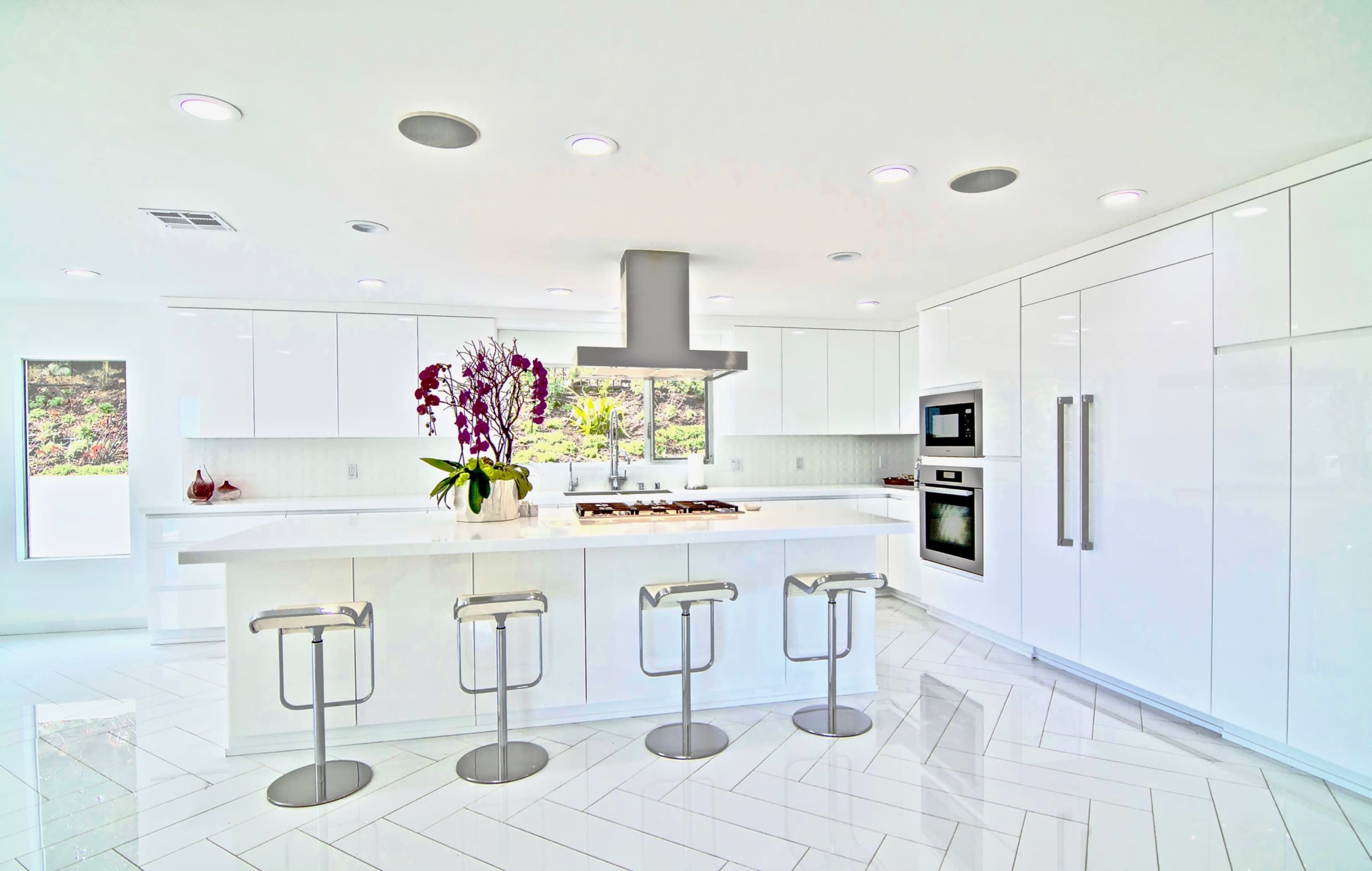 A modern kitchen features white cabinets, a central island with bar stools, and a large window with natural light.