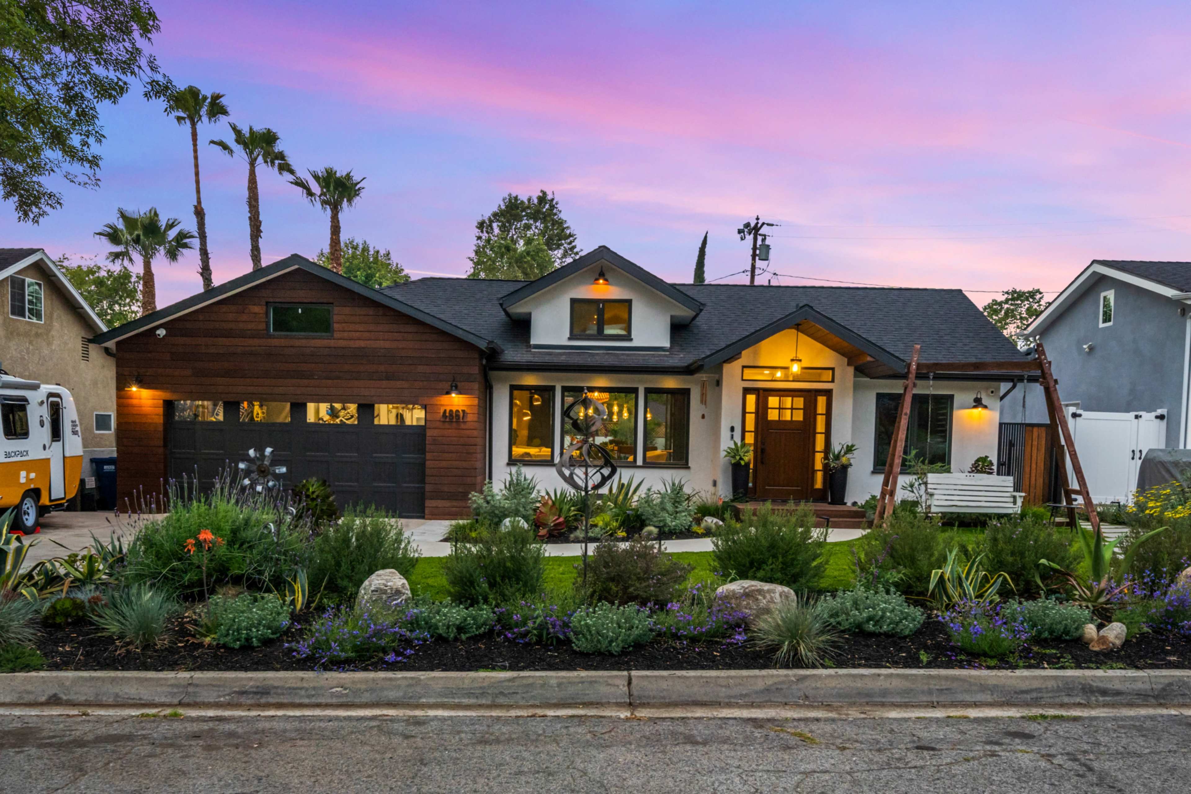 The image shows a modern single-family home with a wooden garage door, surrounded by landscaped gardens, under a colorful sunset sky.