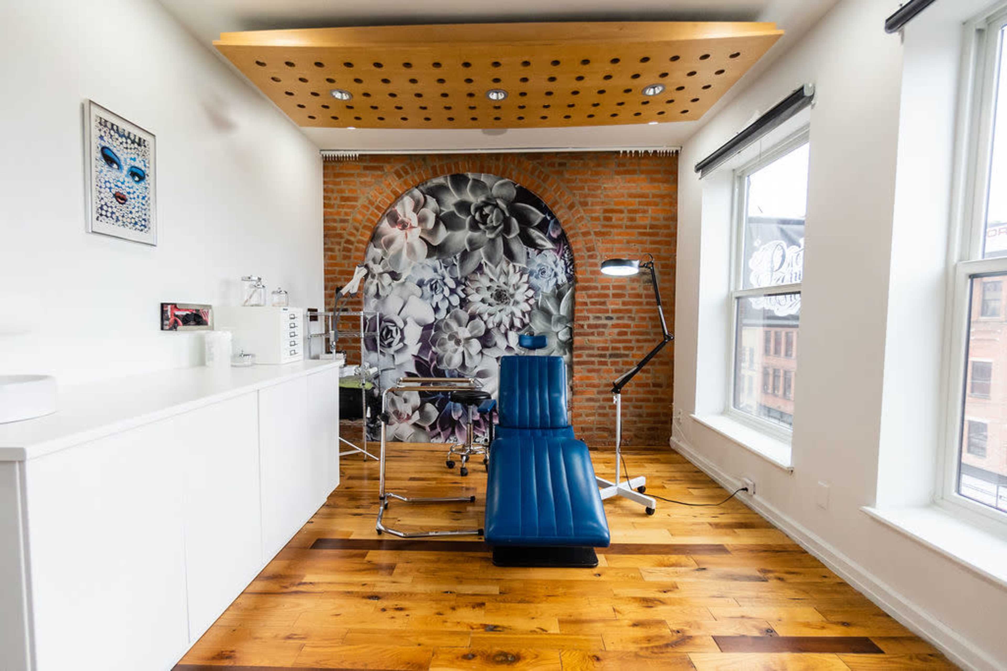 A modern, well-lit treatment room featuring a blue examination chair, a wall adorned with a floral mural, and hardwood flooring.