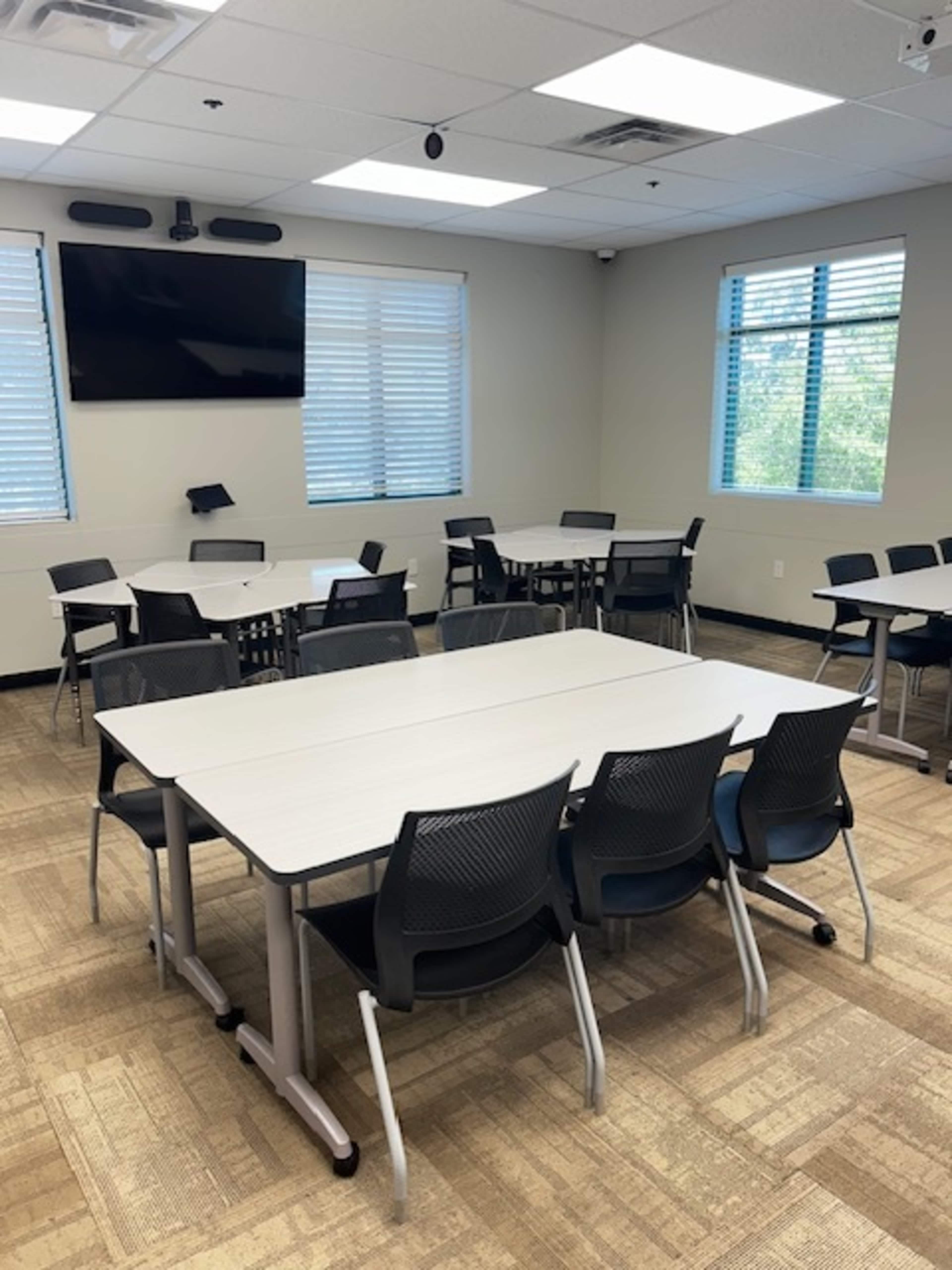 A classroom setting features multiple tables arranged in a layout with black chairs, illuminated by natural light from windows.
