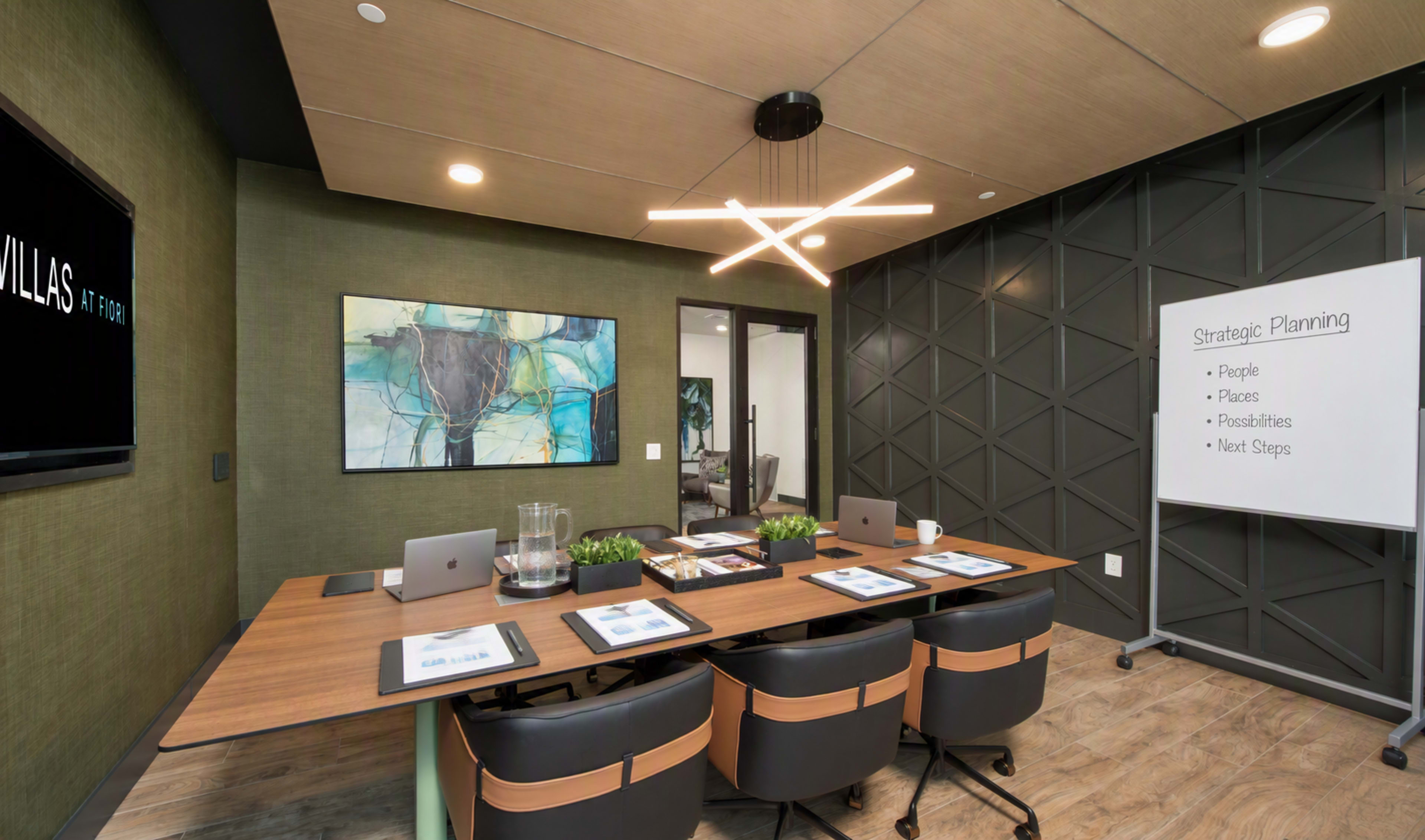 A modern conference room features a large wooden table surrounded by black chairs, with a presentation board, artwork on the wall, and a sleek light fixture overhead.
