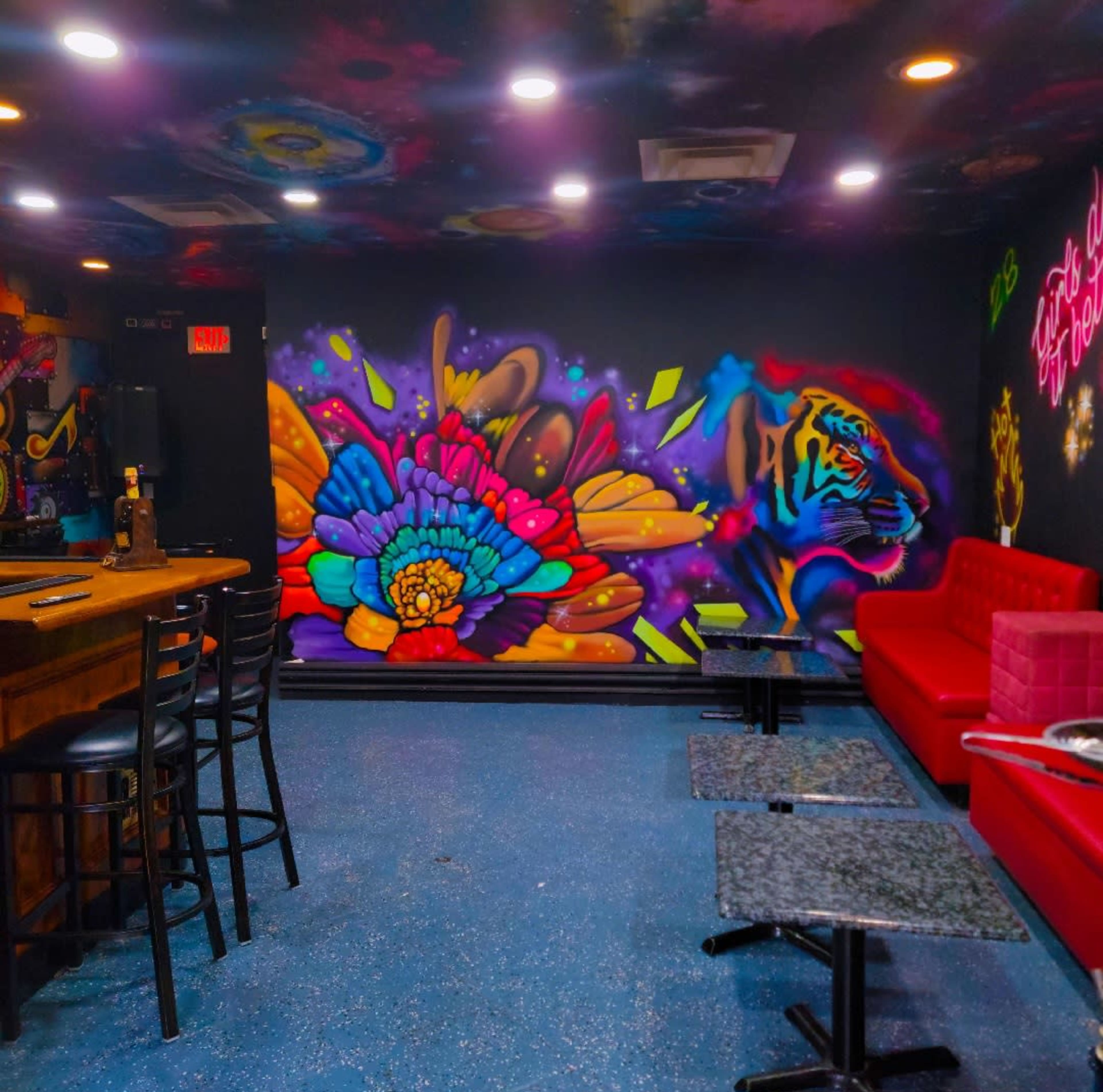 The image shows a vibrant bar area with a colorful mural depicting a tiger and flowers on one wall, and red couches with black tables arranged in the space.