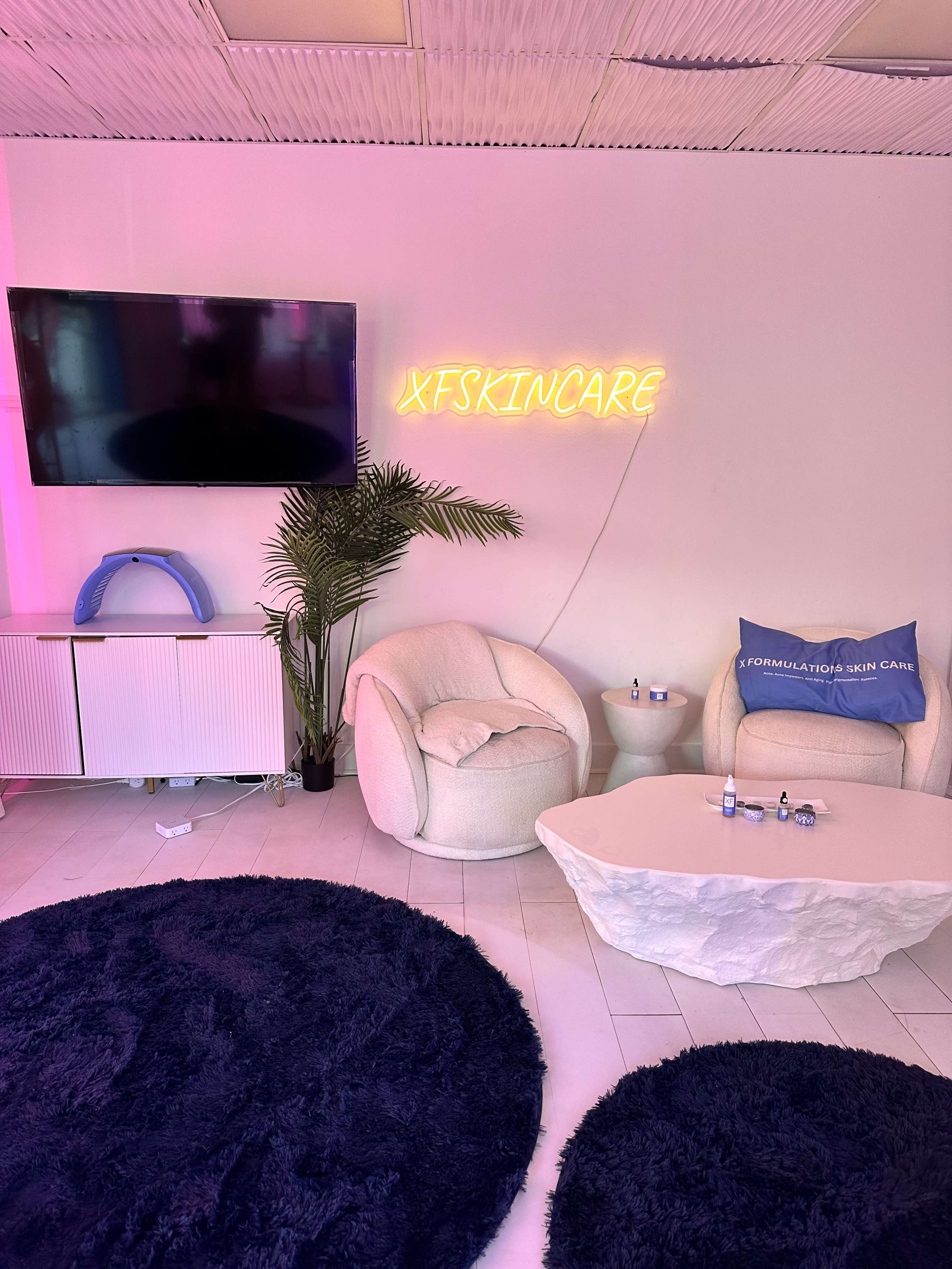 The image shows a modern skincare studio featuring two plush chairs, a television mounted on a pink-lit wall, and a decorative plant next to a coffee table.