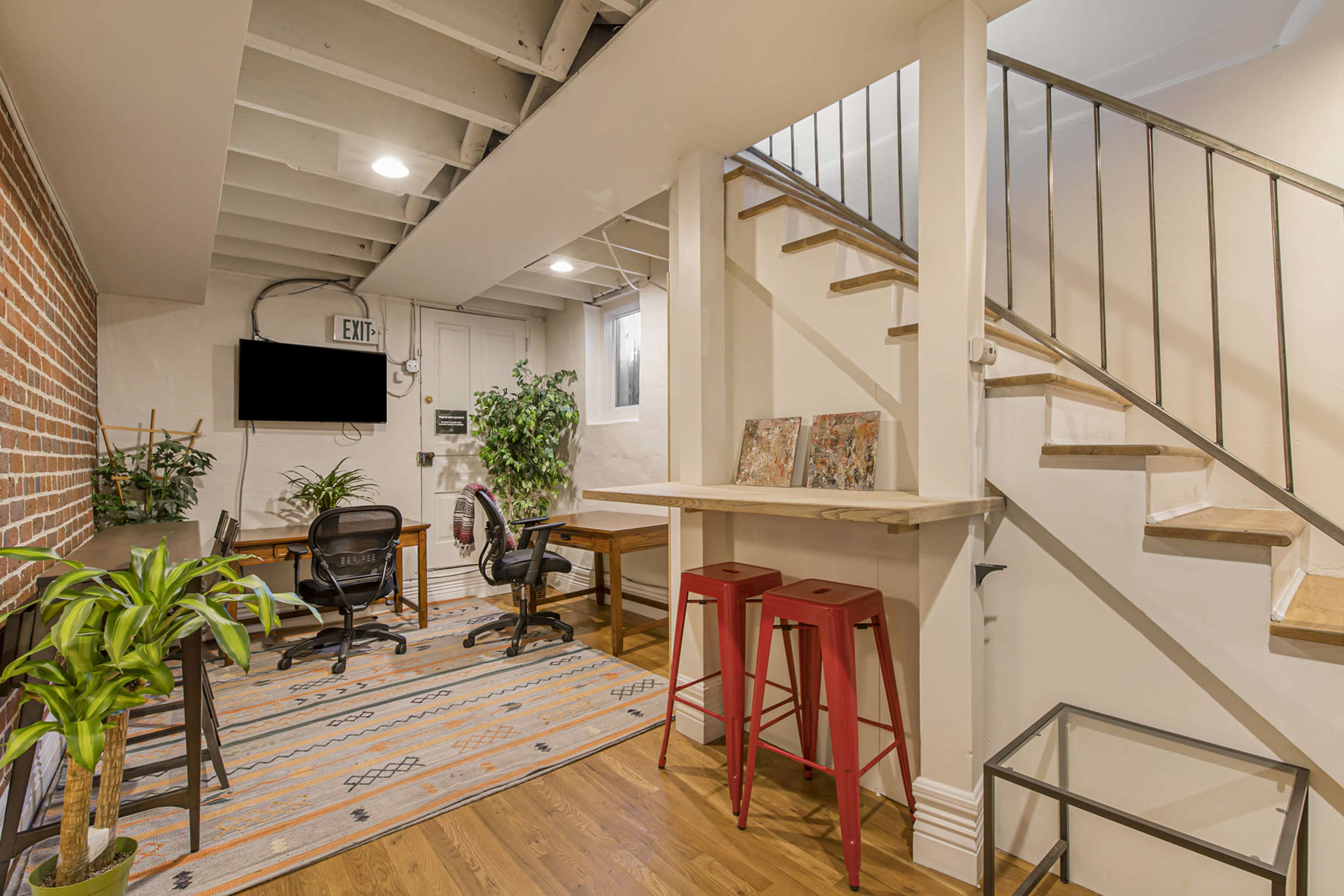 A modern office space featuring a staircase, two workstations, a wall-mounted TV, and greenery.