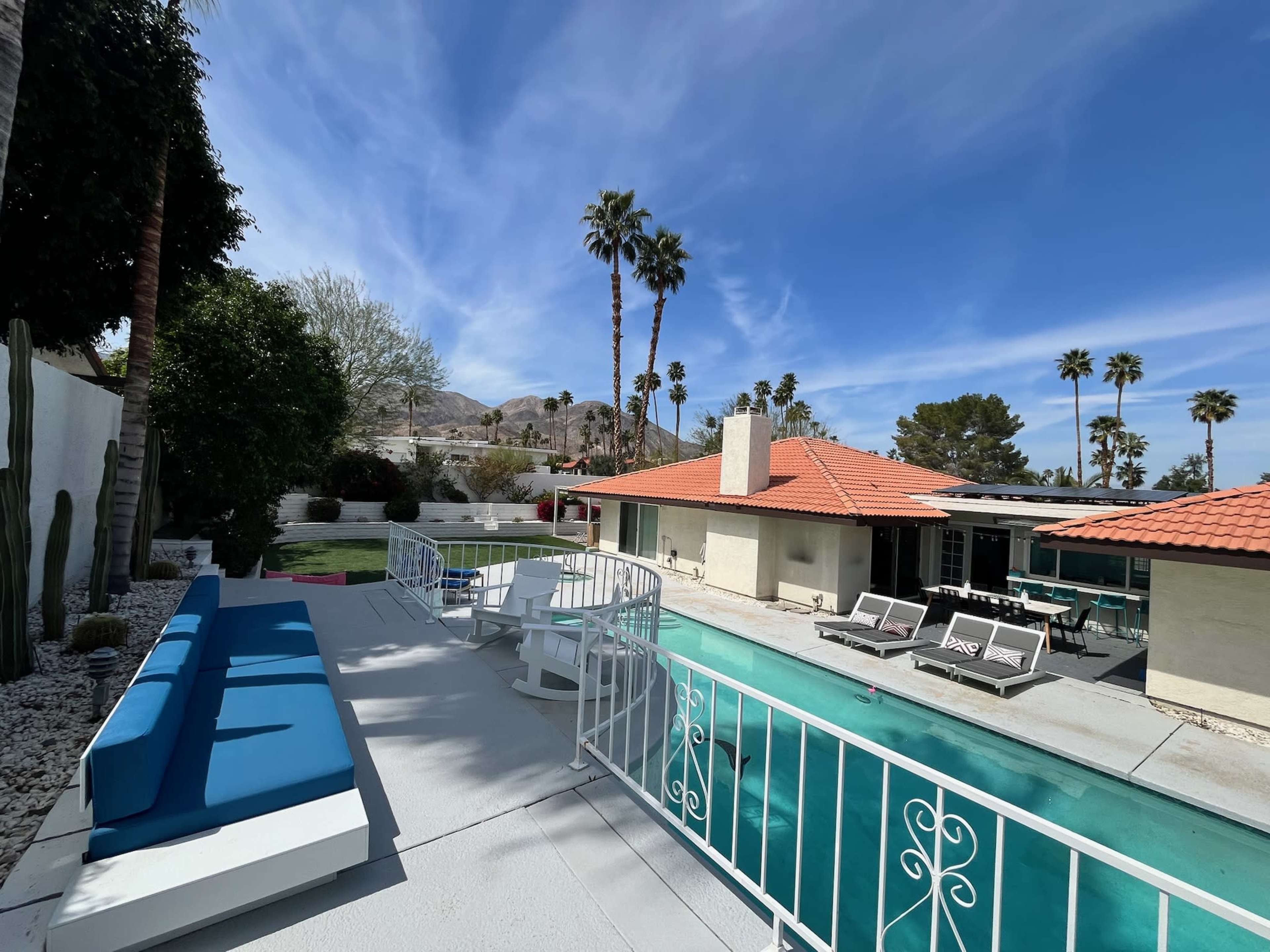 The image shows a patio area with a blue sofa, overlooking a swimming pool surrounded by palm trees and a house with a red-tiled roof.