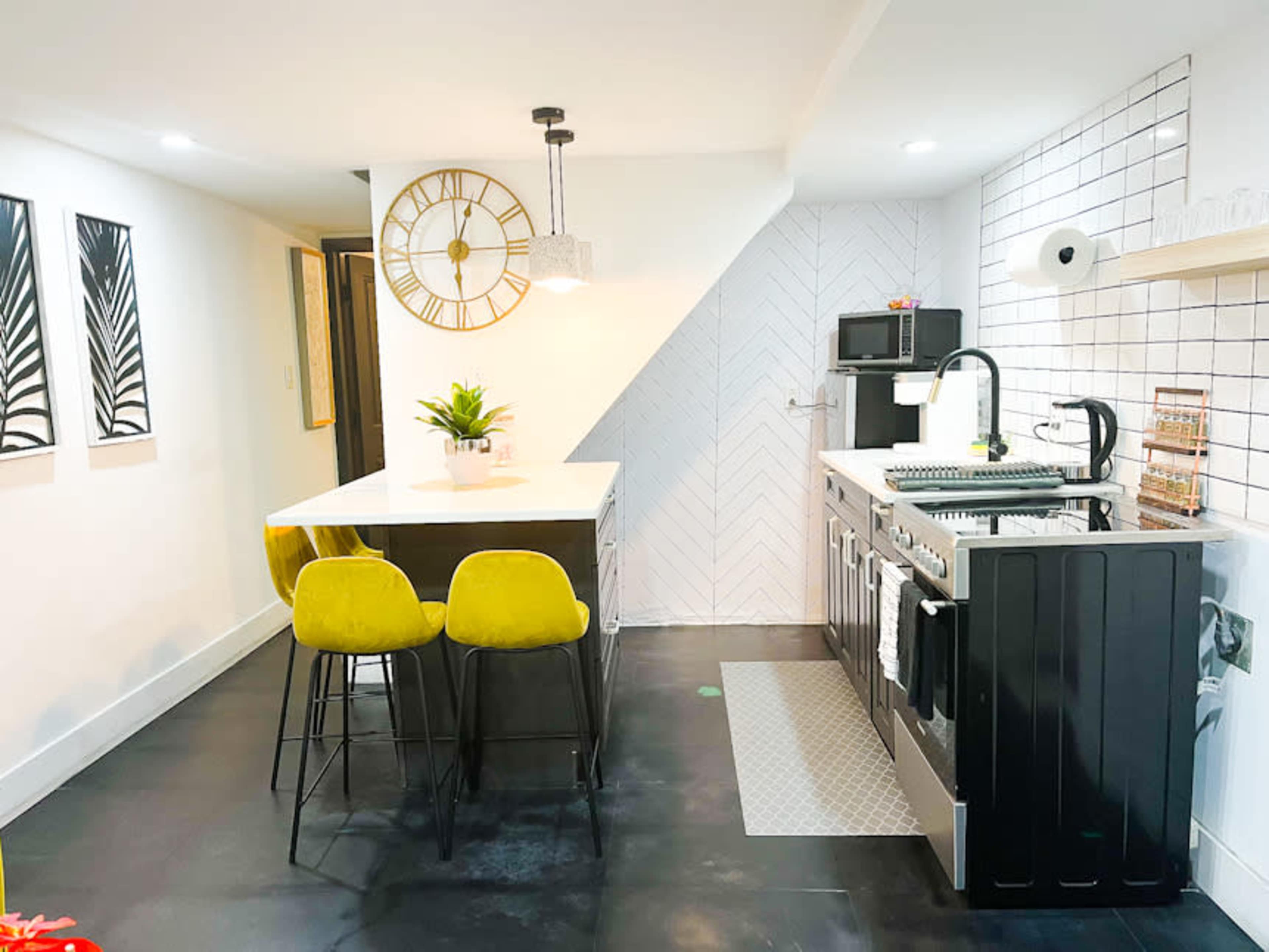 The image shows a modern kitchen with a black and white color scheme, featuring a central island with green stools, stainless steel appliances, and a herringbone-patterned backsplash.