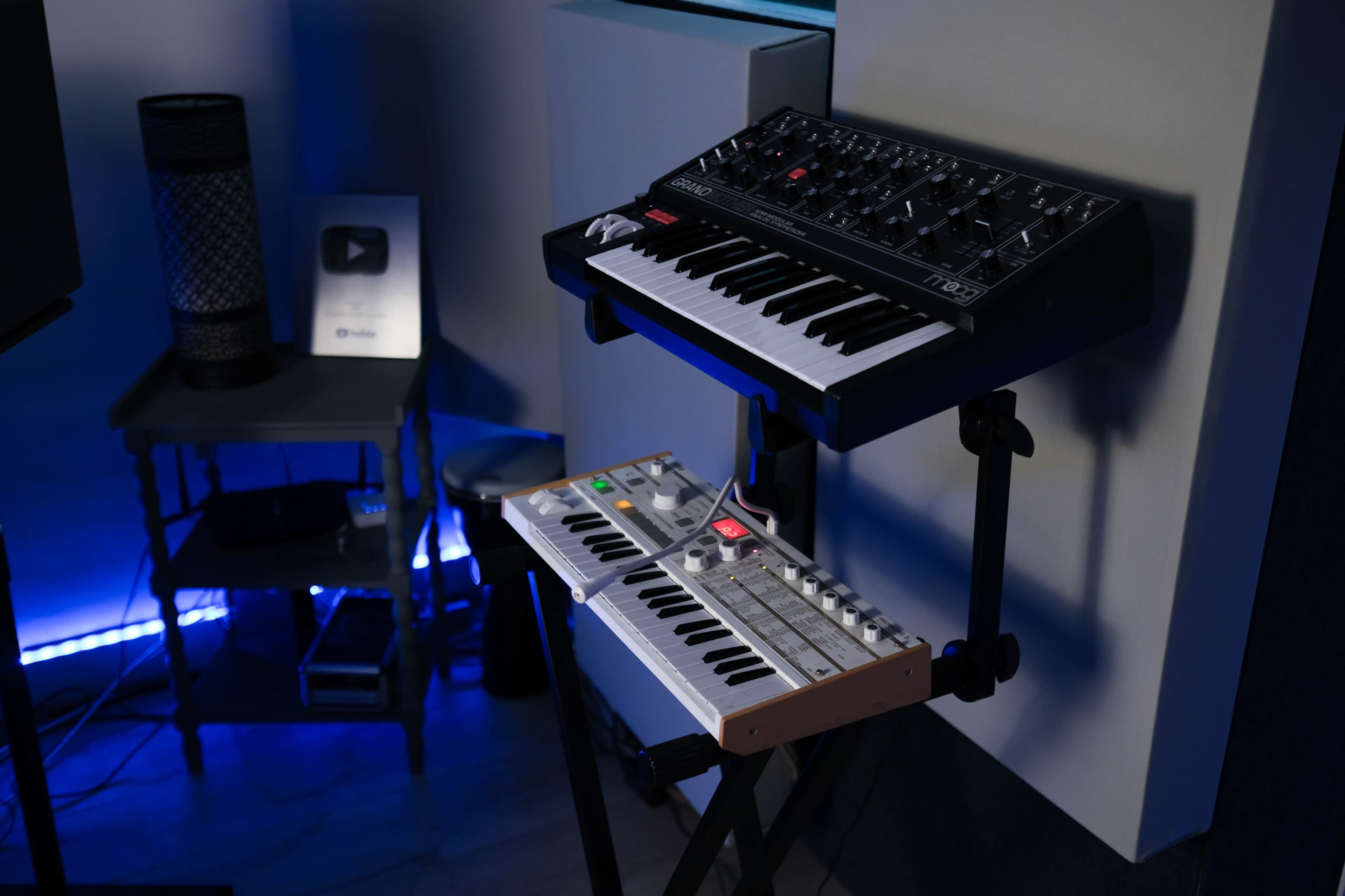 Two synthesizers, one mounted on a stand and the other placed on a lower level, with a softly lit background featuring a blue hue.