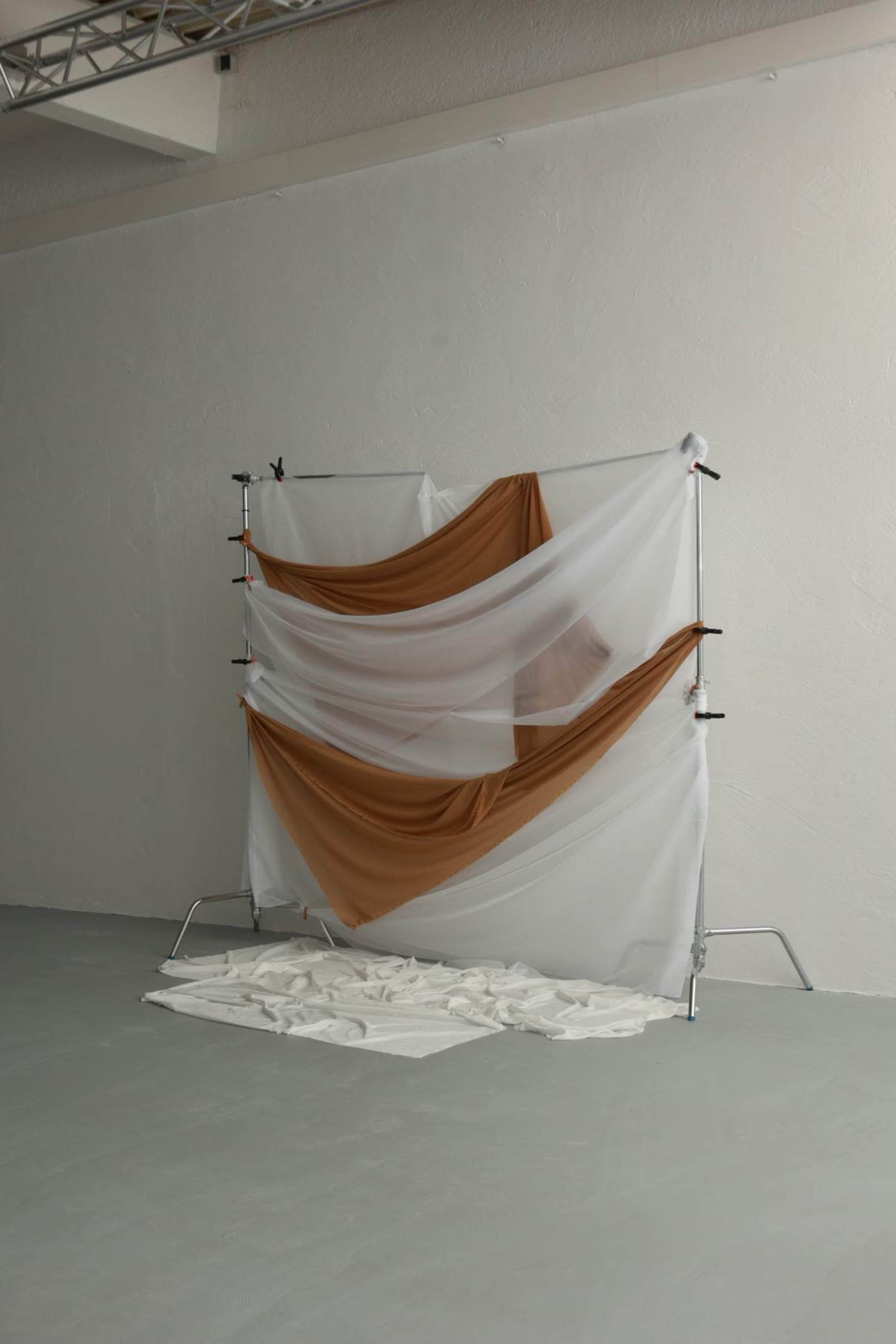 The image shows a minimalist backdrop made of draped fabric in brown and white, supported by a metal frame, set against a plain, light-colored wall.