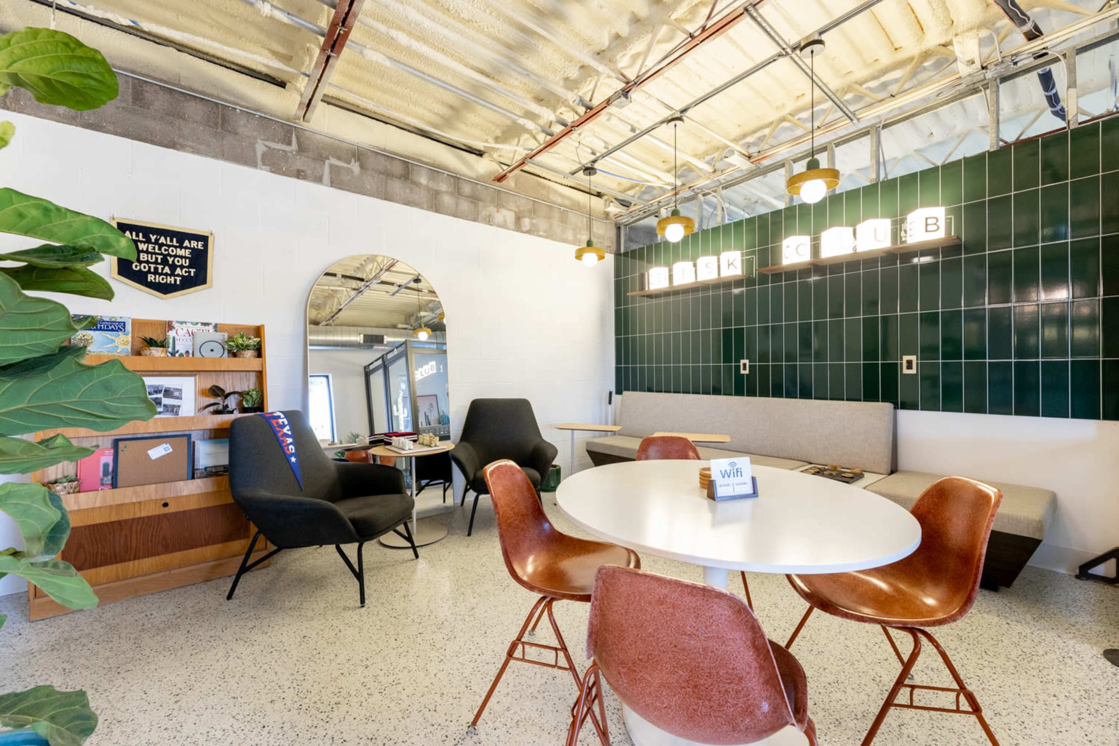 The image shows a modern co-working space with a round table, various seating options, and green tiled walls.