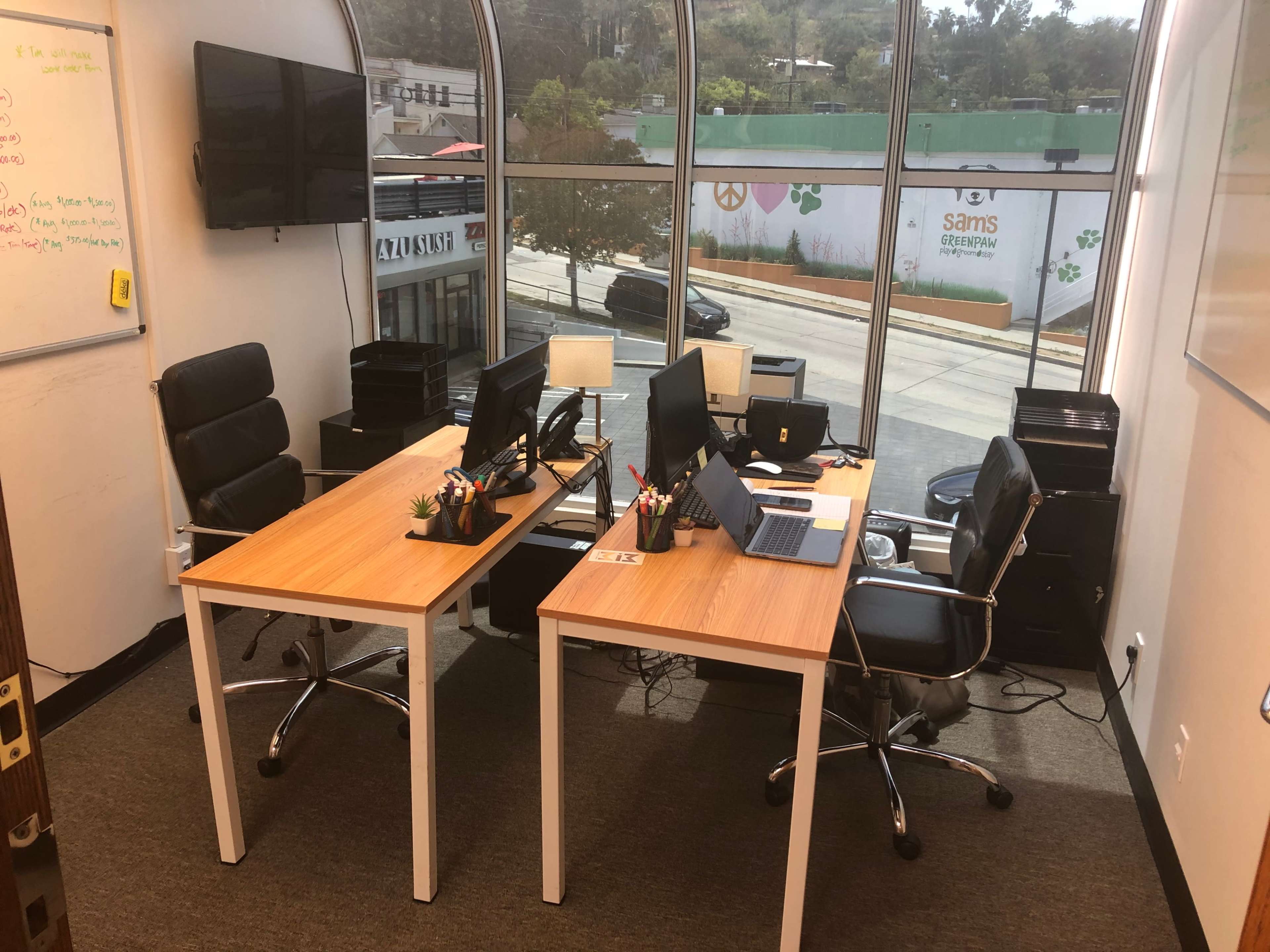 A modern office space with two desks facing each other, each equipped with computers and office supplies, and large windows that overlook a street.