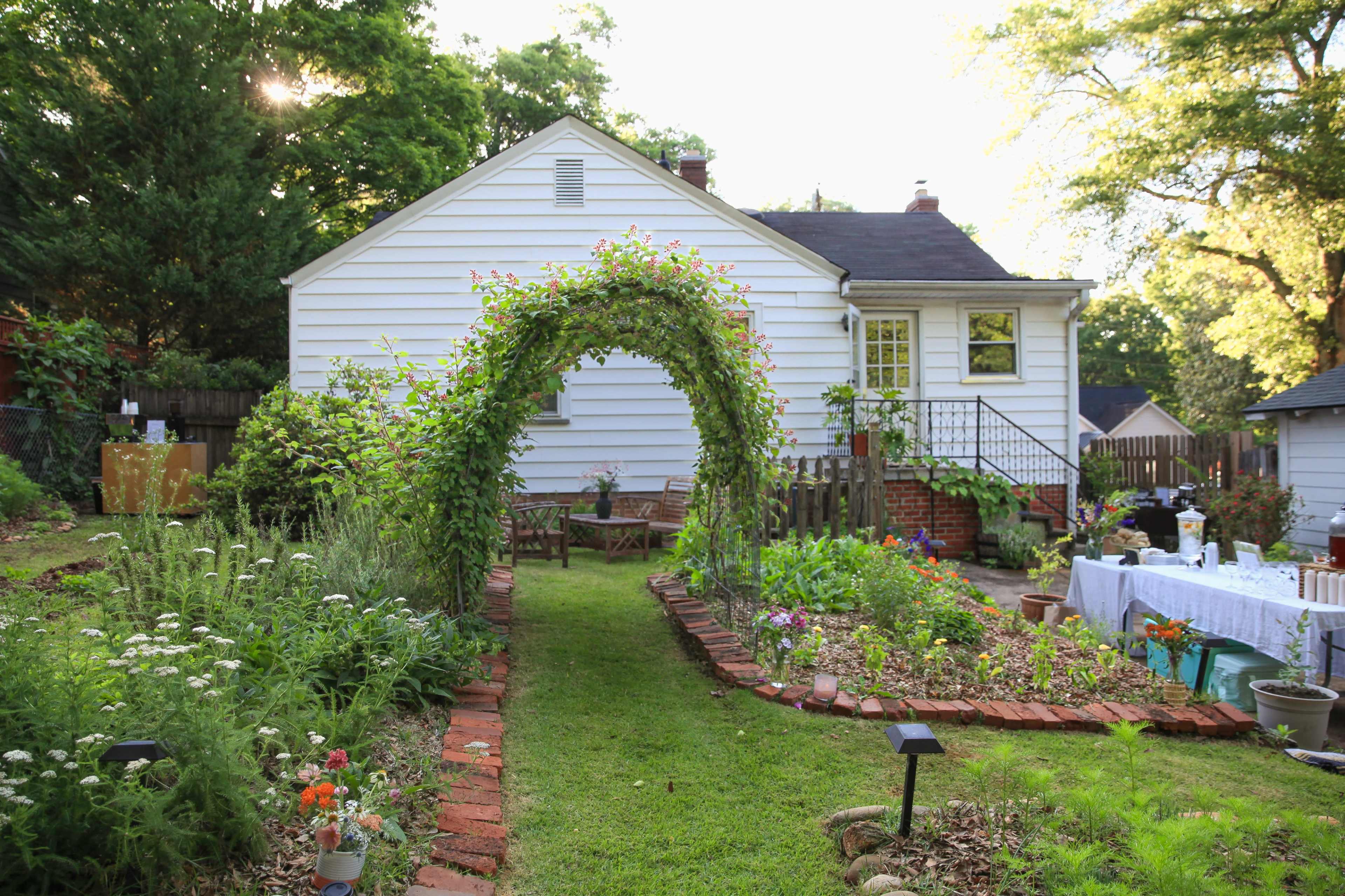 The image shows a well-maintained garden pathway leading to a white house, with a floral arch, flower beds, and outdoor seating.