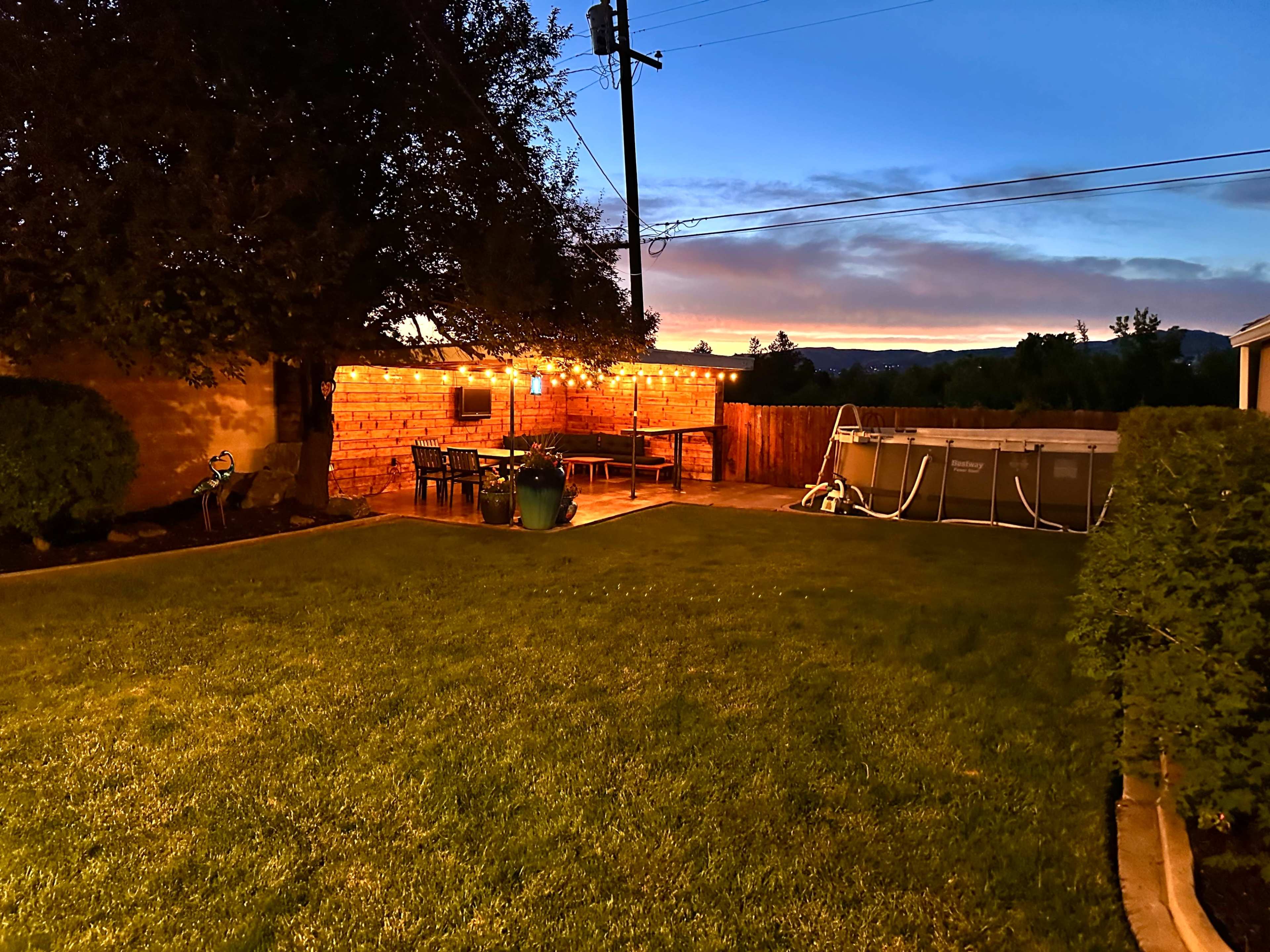 The image shows a backyard with a patio area featuring string lights, a dining set, and a swimming pool, set against a sunset sky.
