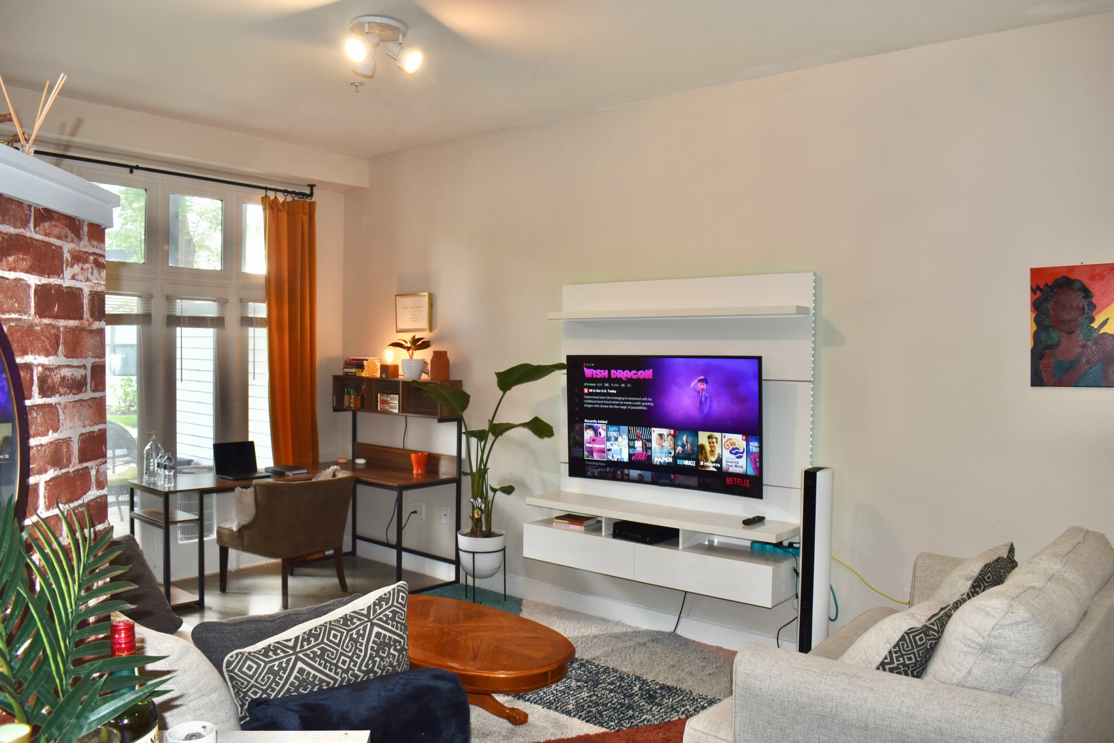 A living room features a mounted television displaying vibrant content, a cozy sofa, a circular coffee table, and a desk setup against a backdrop of large windows.