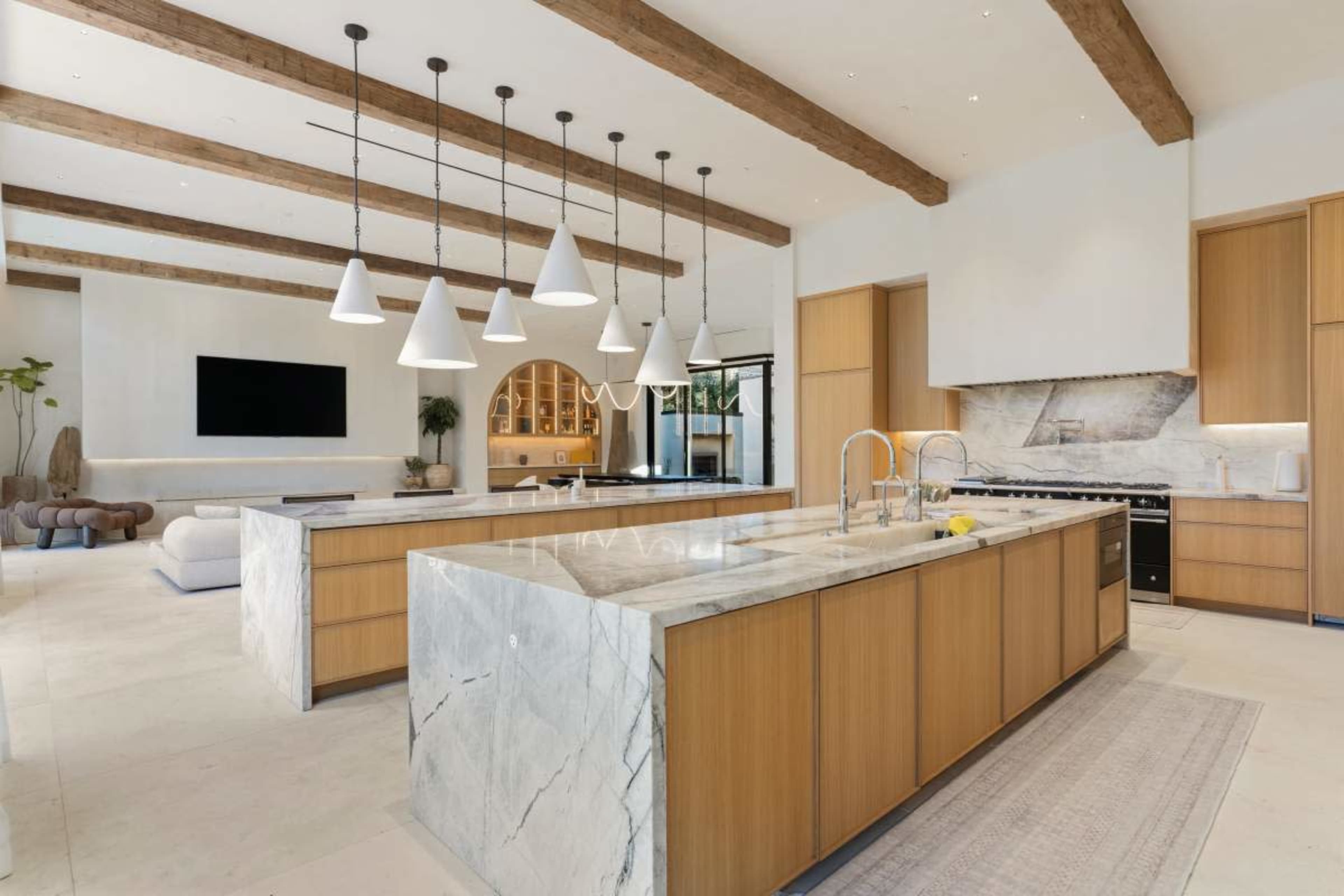 A modern kitchen features large marble countertops, wooden cabinetry, and pendant lighting suspended from the ceiling beams.