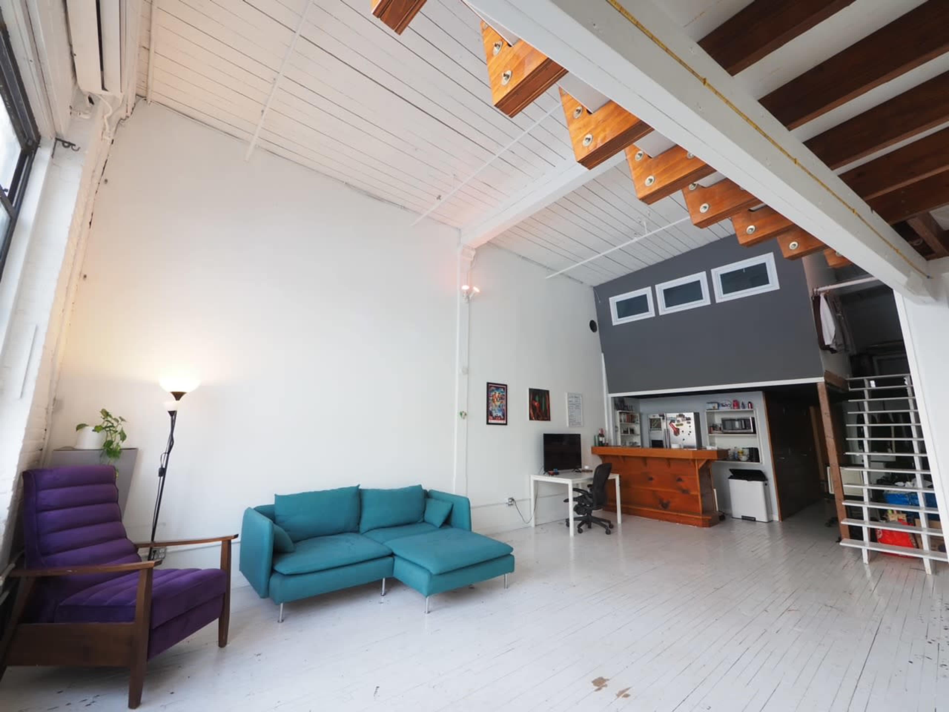 The image shows a spacious living area with a purple armchair, a teal couch, and wooden stairs leading to a lofted space.